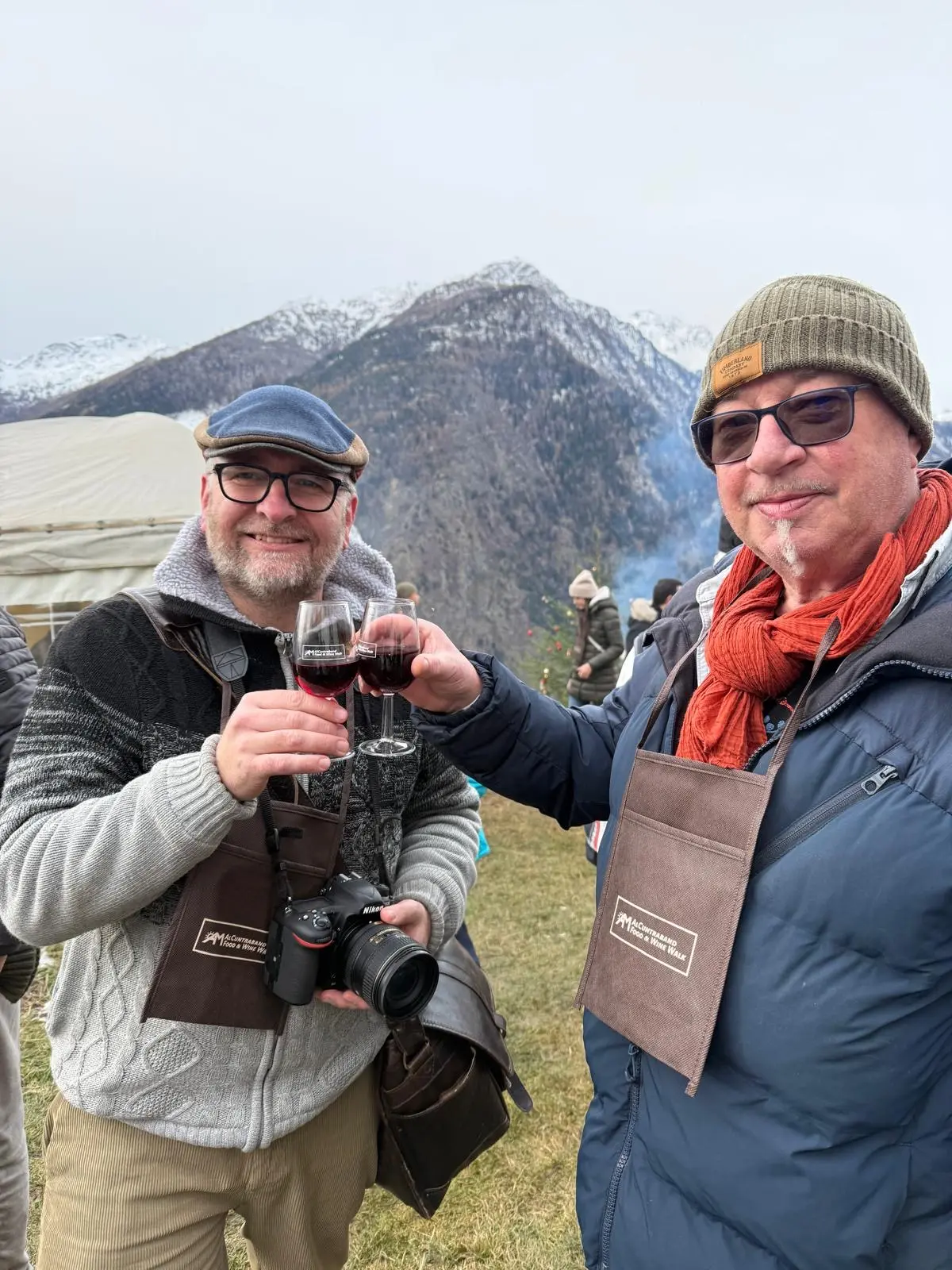 Due uomini sorridenti, ciascuno con un bicchiere di vino rosso, brindano insieme. Sullo sfondo si vedono montagne coperte di neve e un cielo nuvol