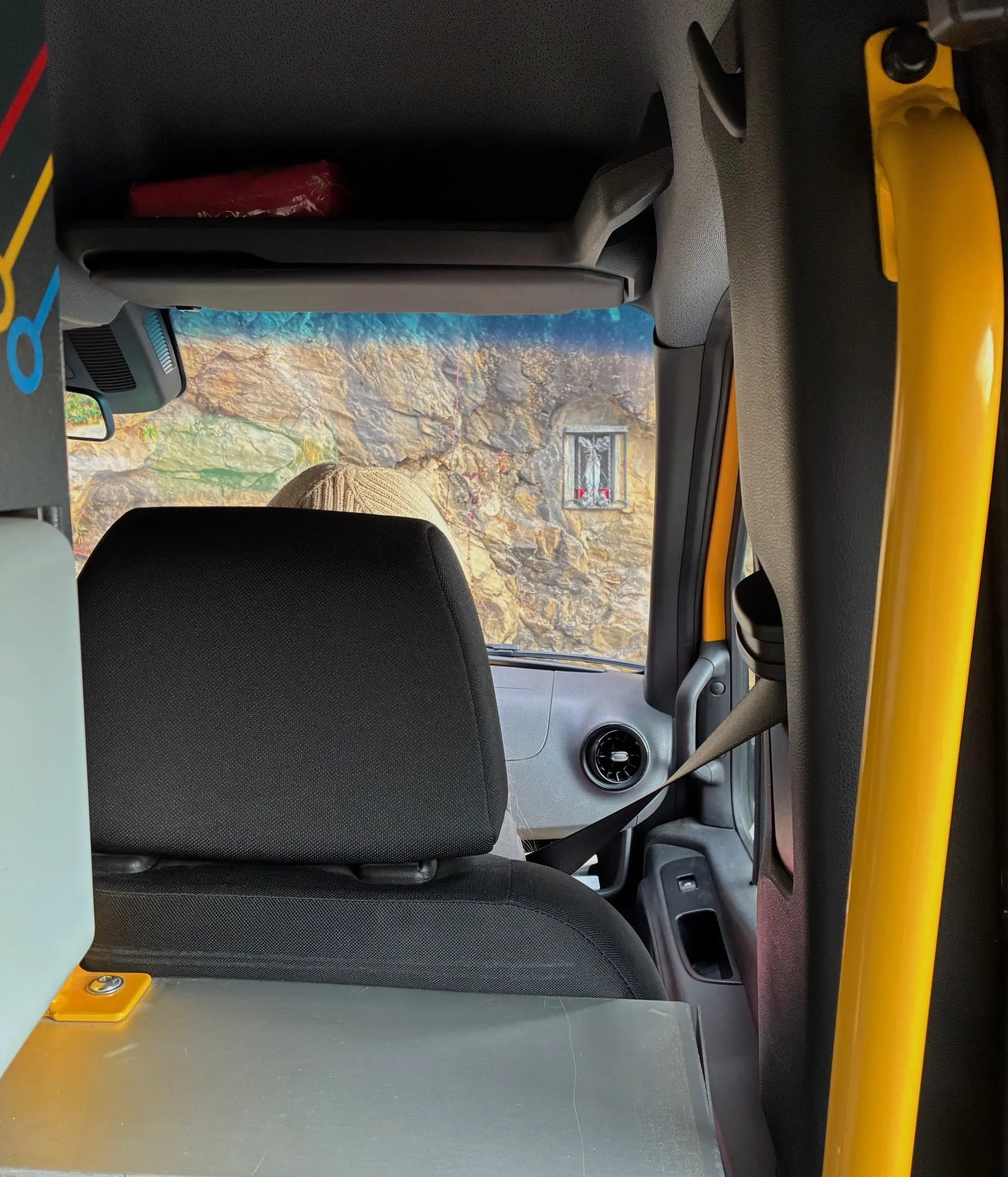 Interno di un veicolo con una vista attraverso il parabrezza. Un sedile è visibile sul retro, con un cappello in primo piano e un paesaggio roccioso che