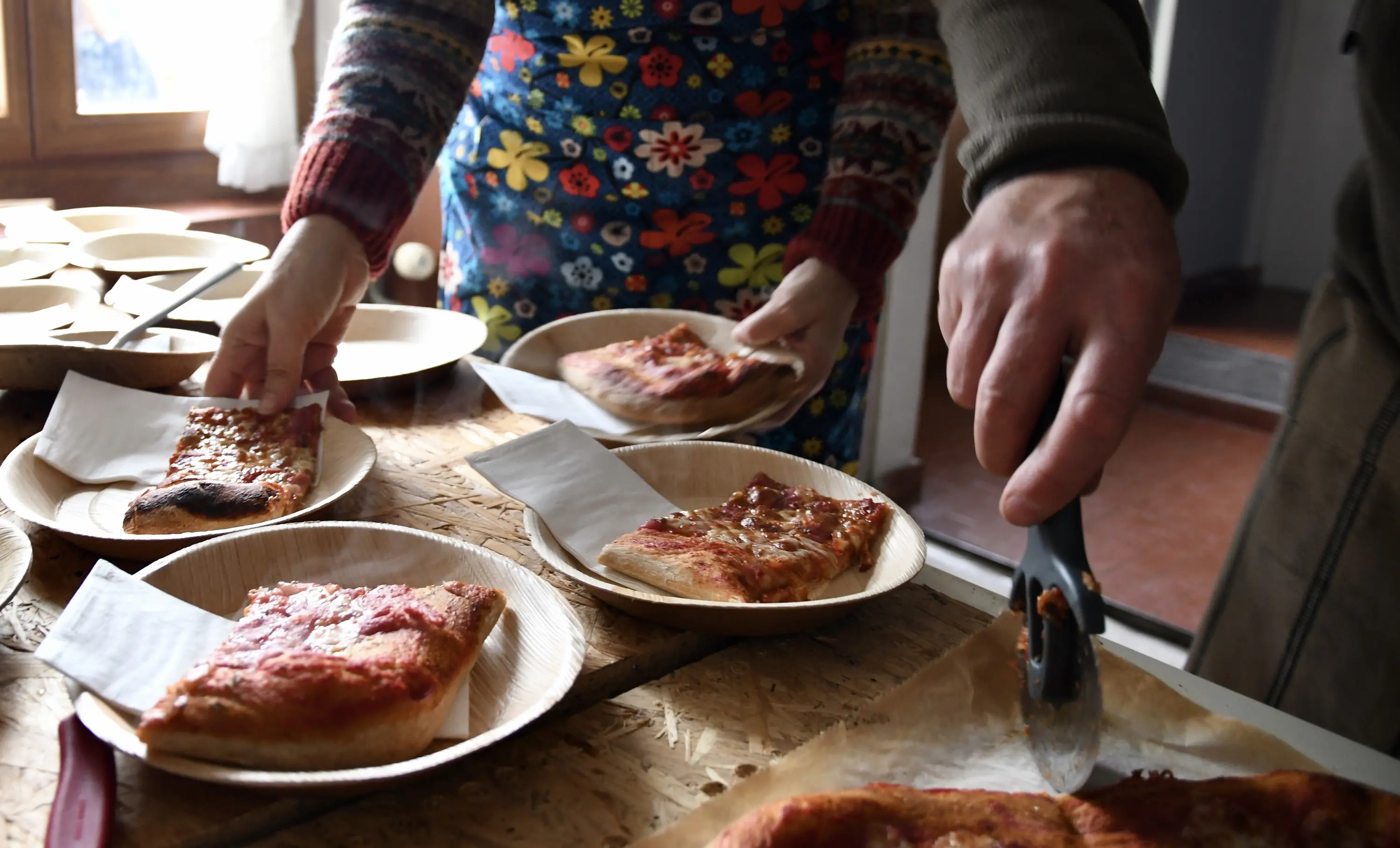 Persone servono fette di pizza su piatti di legno, con tovaglioli bianchi. Una persona indossa un grembiule colorato mentre l'altra utilizza una forche