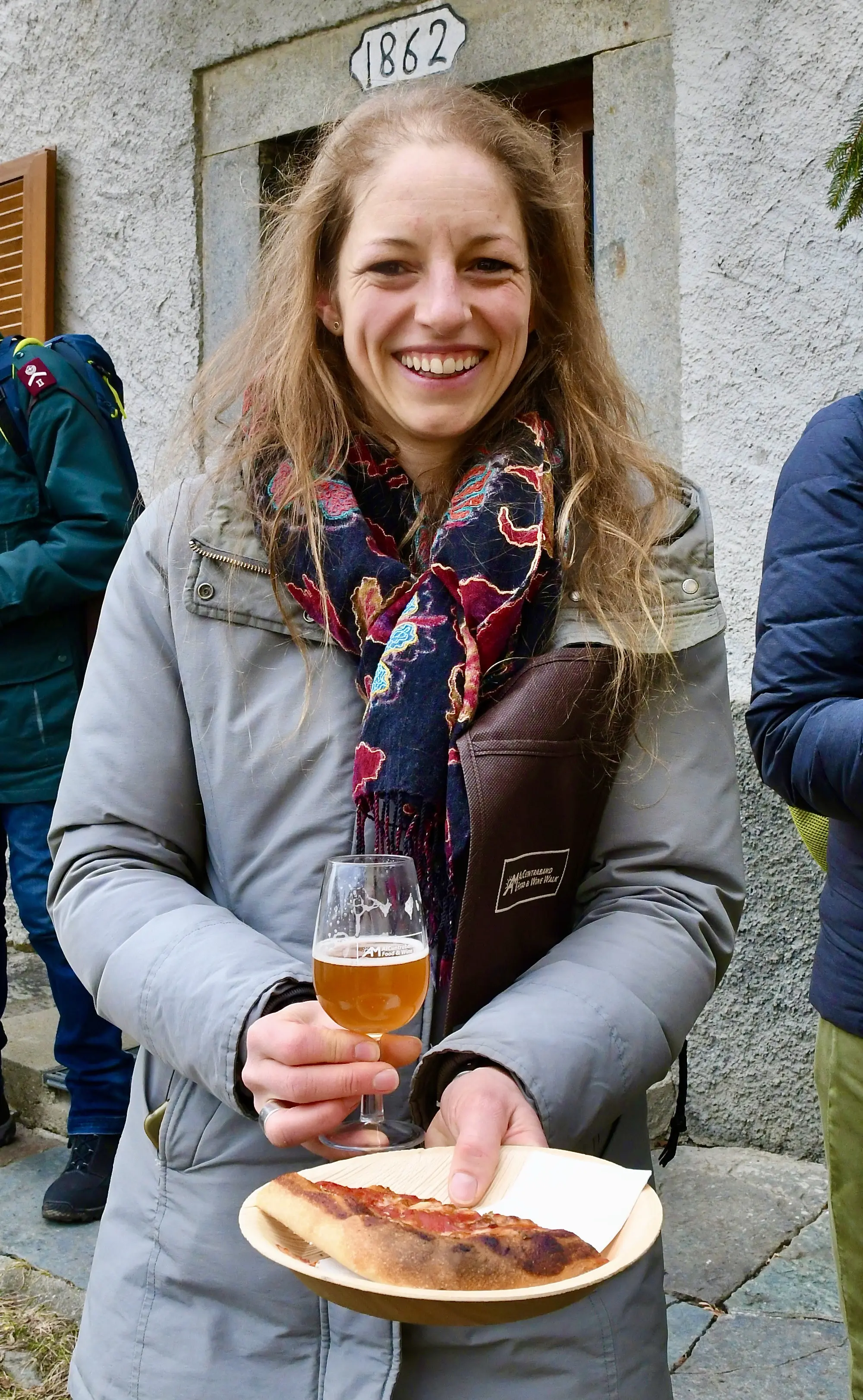 Una giovane donna sorride mentre tiene in mano un bicchiere di birra e un piatto di cibo. Indossa un cappotto grigio e una sciarpa colorata.