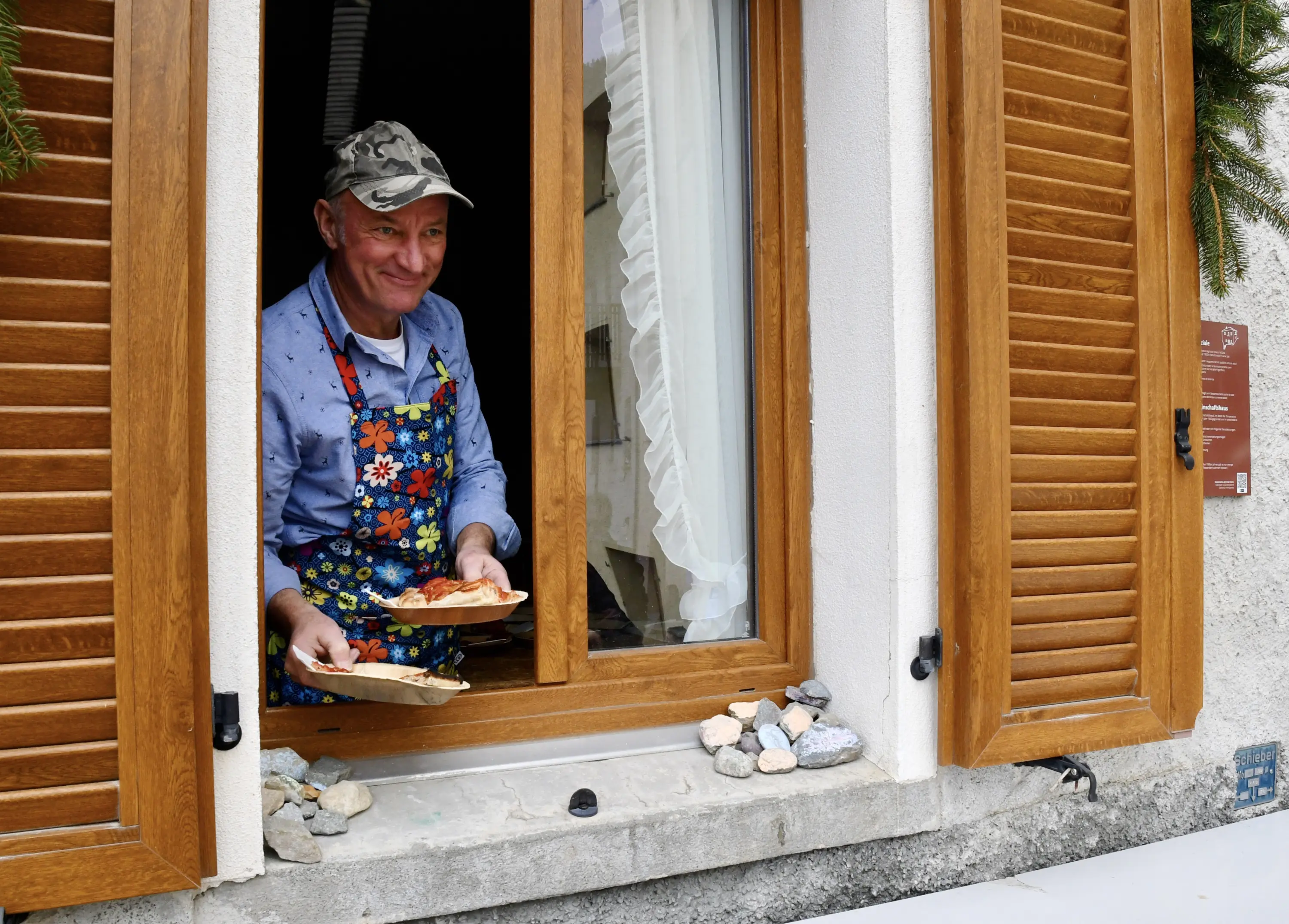 Un uomo sorridente indossa un grembiule colorato e tiene un piatto con del cibo mentre si affaccia da una finestra con persiane di legno.