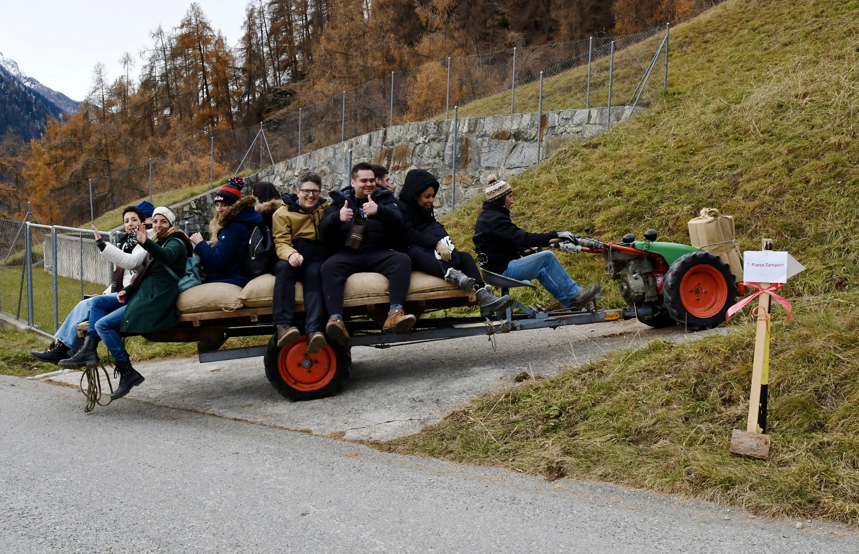 Un gruppo di sette persone sedute su un rimorchio trainato da un trattore, sorridendo e facendo segni di approvazione. Sullo sfondo si vedono alberi