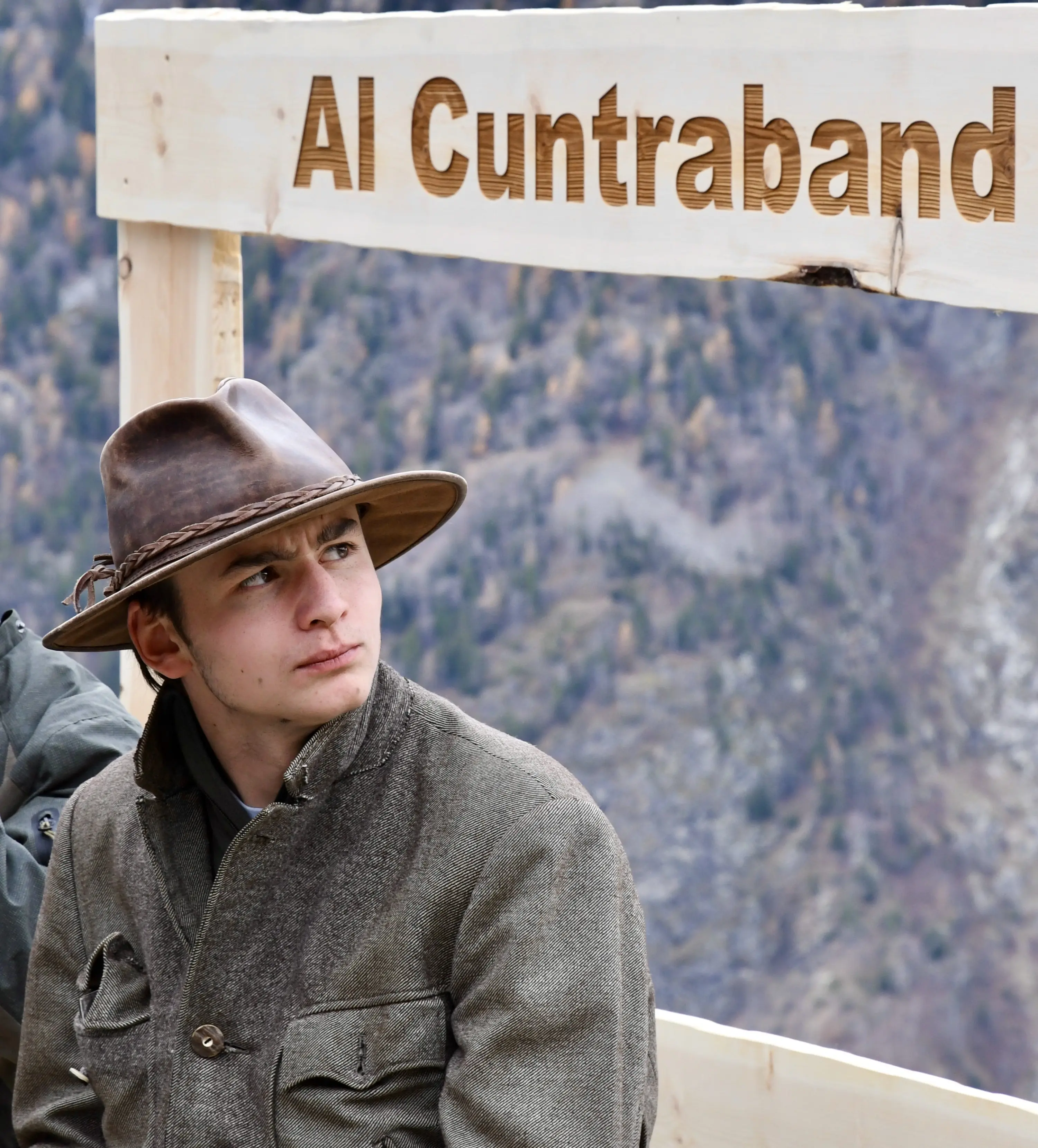 Un giovane con un cappello di feltro siede accanto a un cartello in legno che dice "Al Cuntraband". Sullo sfondo si vedono alberi e montagne.