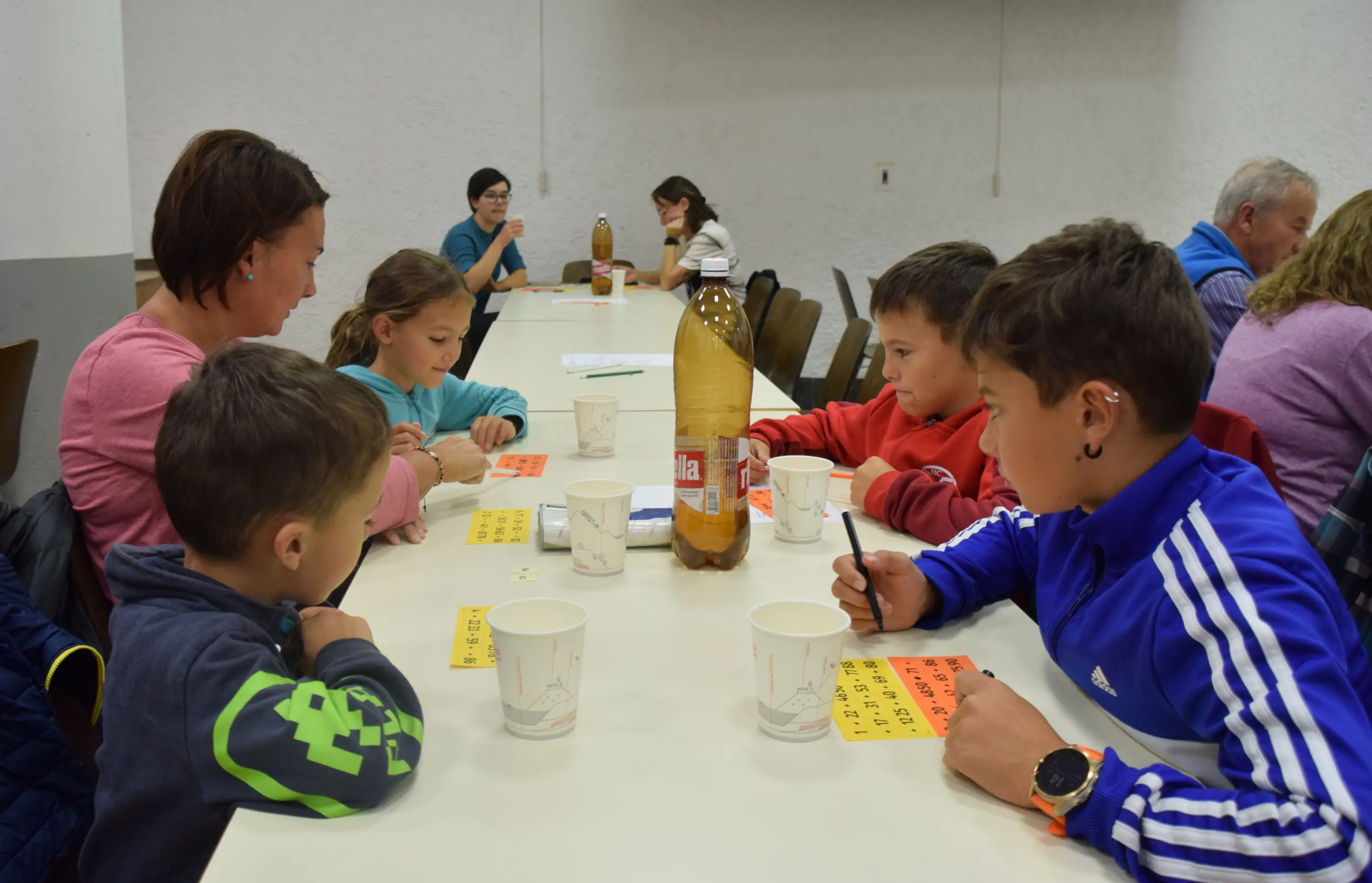 Un gruppo di adulti e bambini seduti a un tavolo, impegnati in un'attività creativa. Alcuni stanno scrivendo su fogli gialli, mentre altri osservano. Al centro del tavolo è presente una bottiglia di plastica e ci sono anche bicchieri di carta.