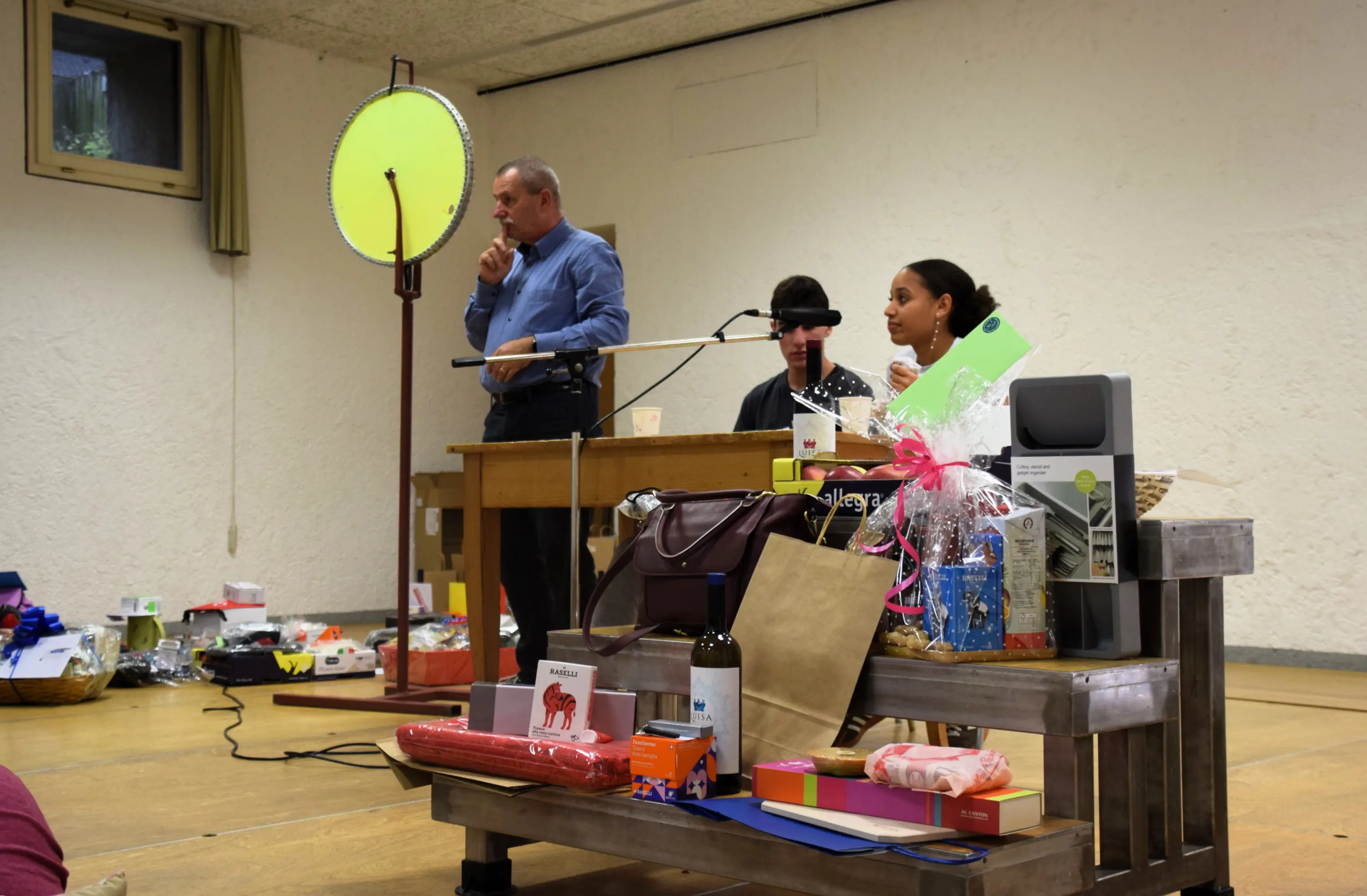 Un palco con un uomo che parla al microfono, una donna seduta accanto a lui e una persona con un copricapo. Davanti ci sono vari regali e oggetti in esposizione su tavoli di legno. Una luce circolare green è appesa alle pareti.