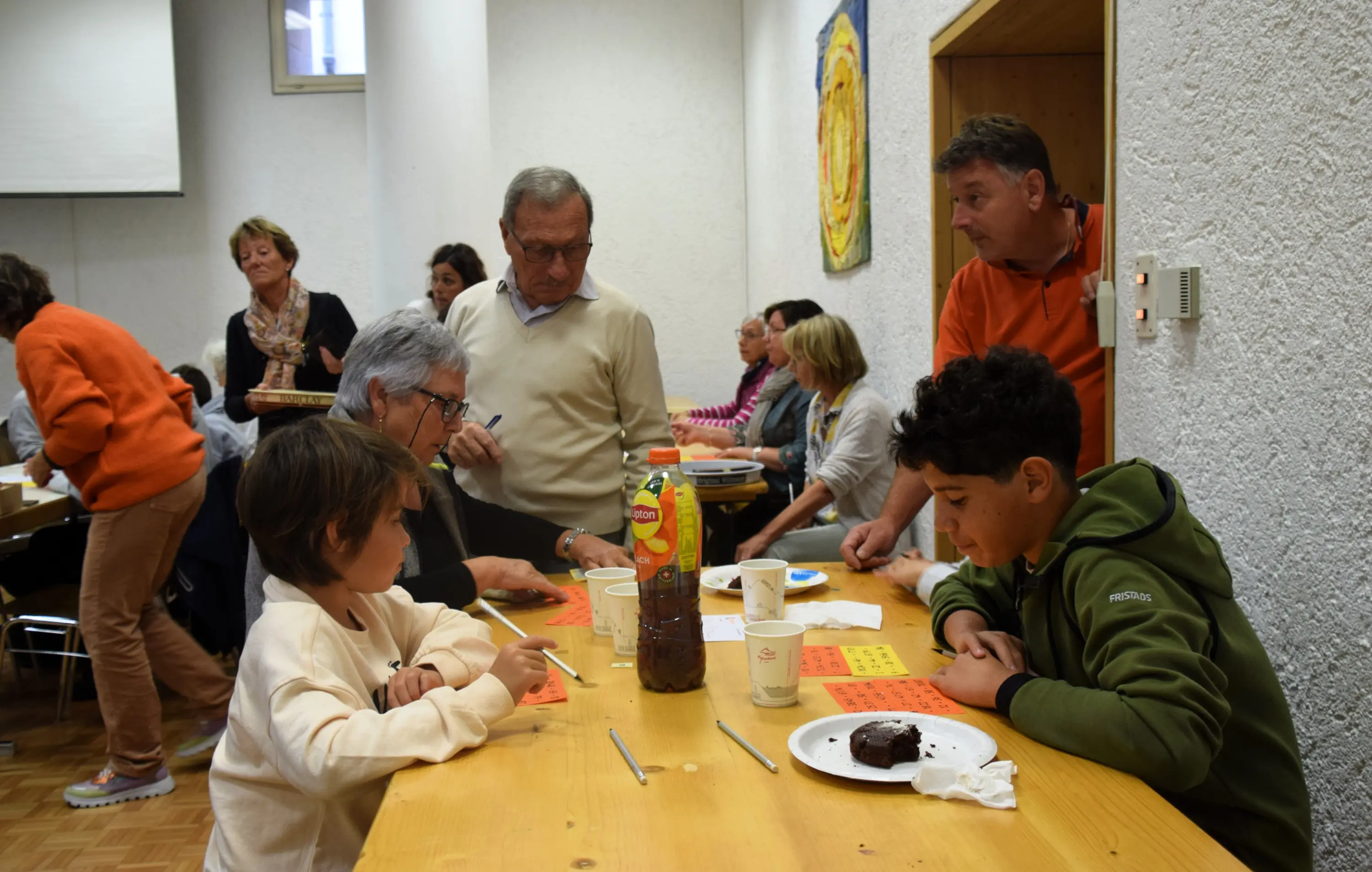Un gruppo di persone si riunisce in una sala, alcuni seduti a un tavolo mentre altri si muovono. Due bambini sono concentrati su un'attività, con una torta al cioccolato e bevande sul tavolo. Gli adulti interagiscono tra di loro e con i bambini, creando un'atmosfera conviviale.