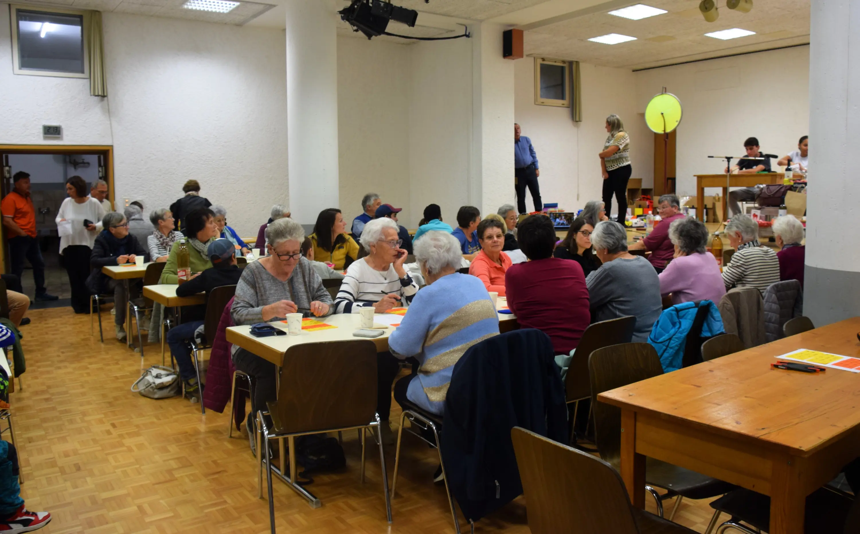 Un gruppo di persone anziane si riunisce in una sala comunale, sedute ai tavoli, mentre alcune interagiscono tra loro. Sullo sfondo, un palco con una persona che parla e un'altra che osserva. L'atmosfera è vivace e sociale.