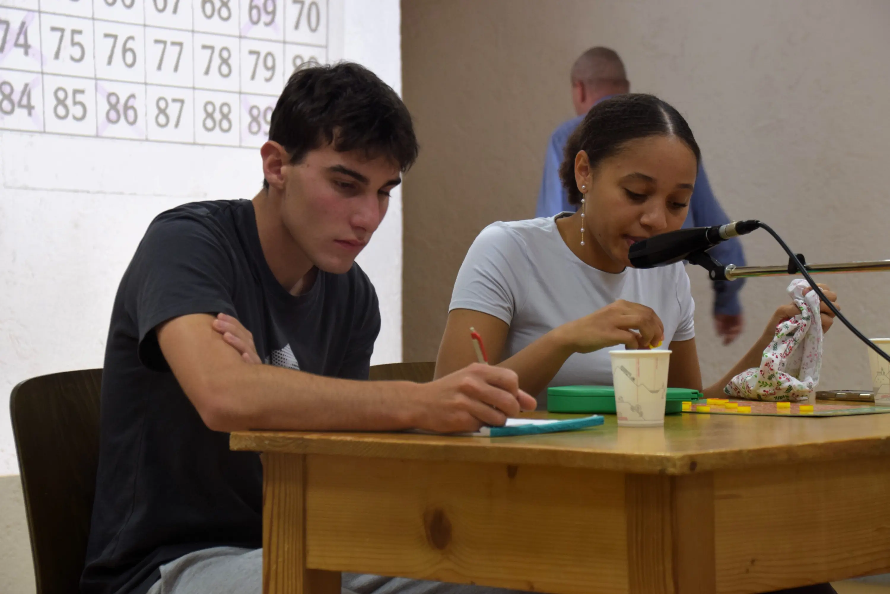 Due giovani stanno seduti a un tavolo. Un ragazzo scrive su un quaderno, mentre una ragazza parla al microfono, tenendo in mano un bicchierino di plastica. Sullo sfondo, un'immagine di numeri è visibile su un muro.