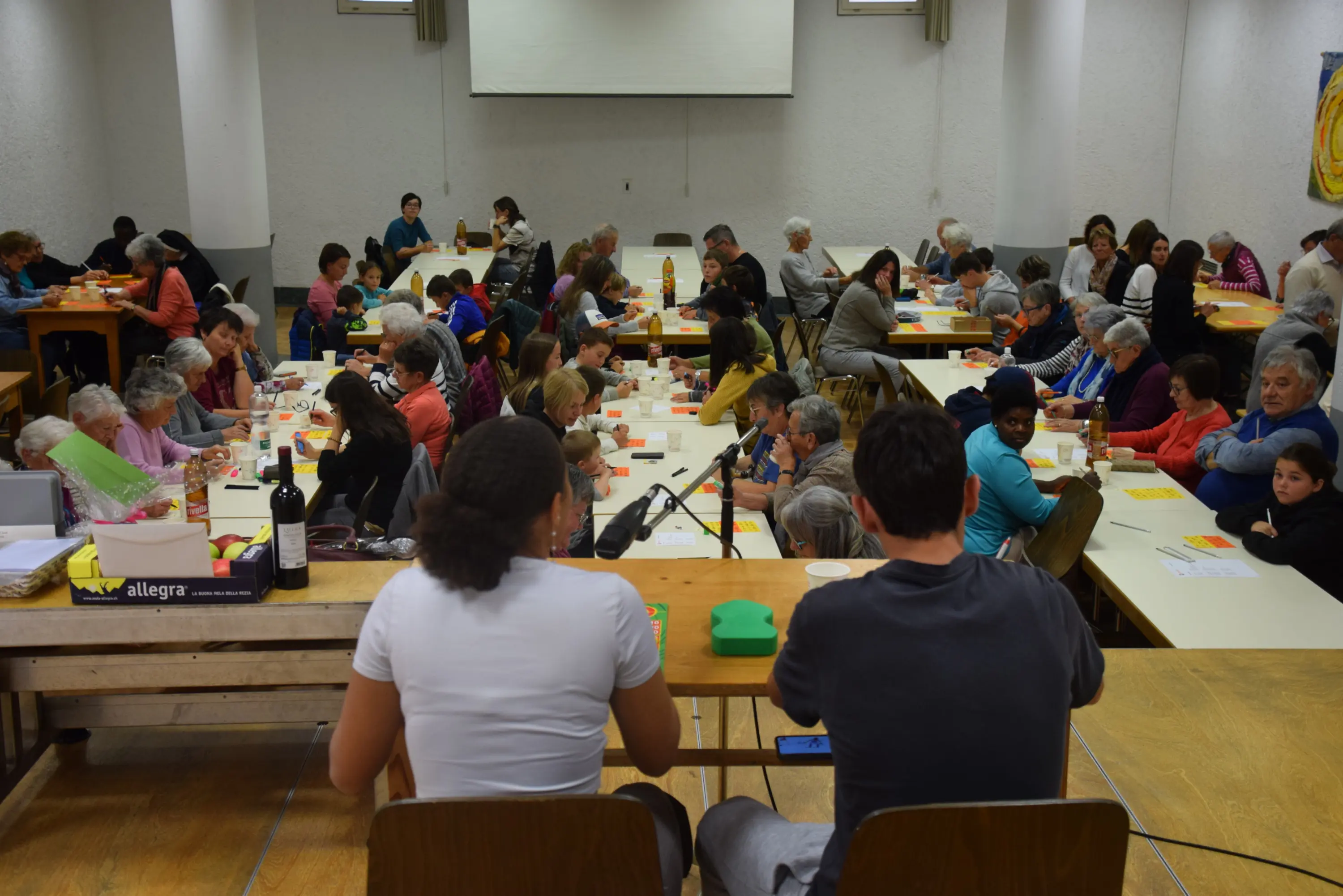 Una sala gremita di persone sedute a tavoli, impegnate in attività manuali. Alcuni bambini e adulti stanno creando lavori artistici, mentre altri chiacchierano. In primo piano, due giovani sono seduti e osservano l'evento.