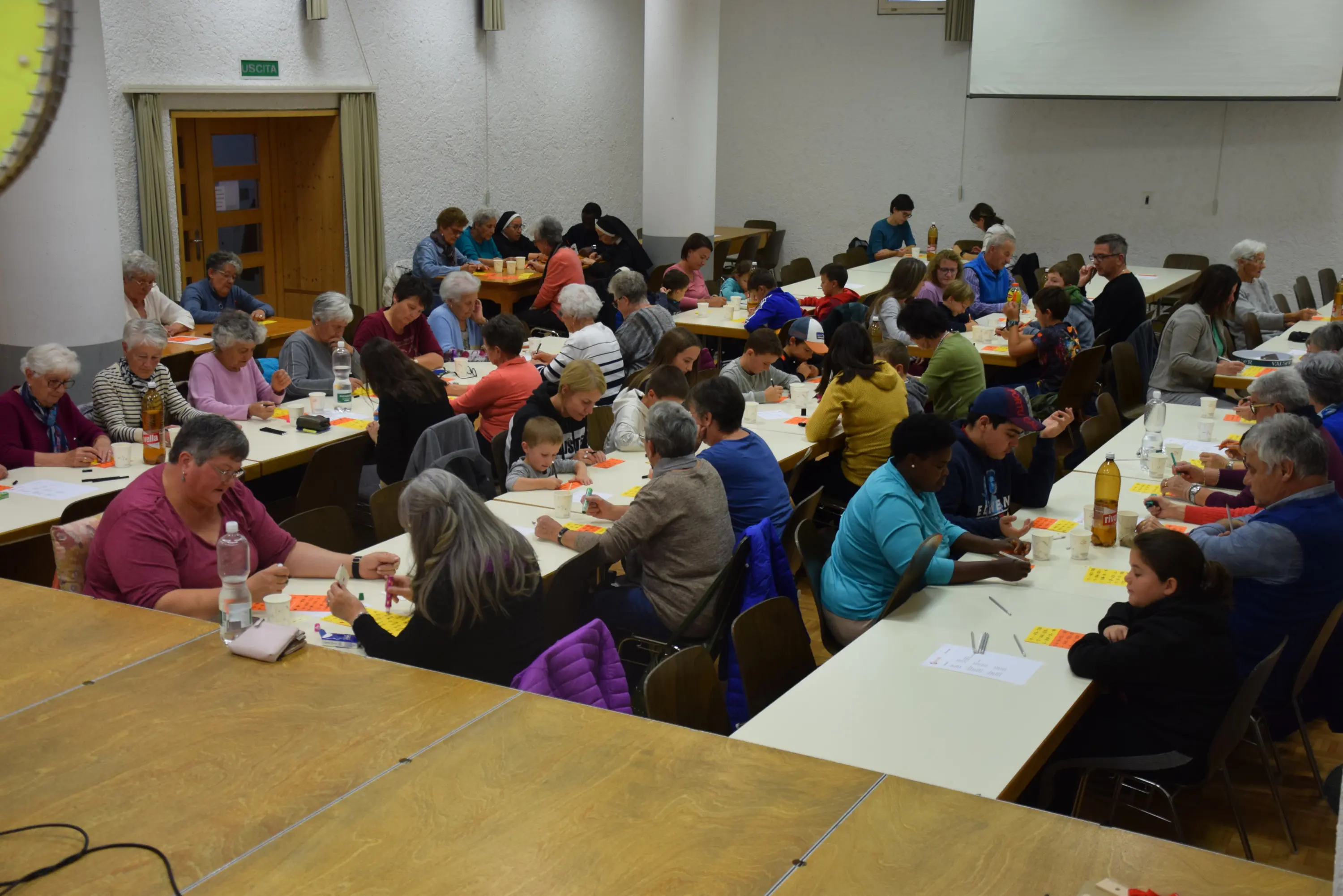 Un gruppo numeroso di persone, tra cui anziani e bambini, è seduto a tavoli in una sala. Stanno giocando a tombola, con cartelle e segnapunti. L'atmosfera è vivace e conviviale.
