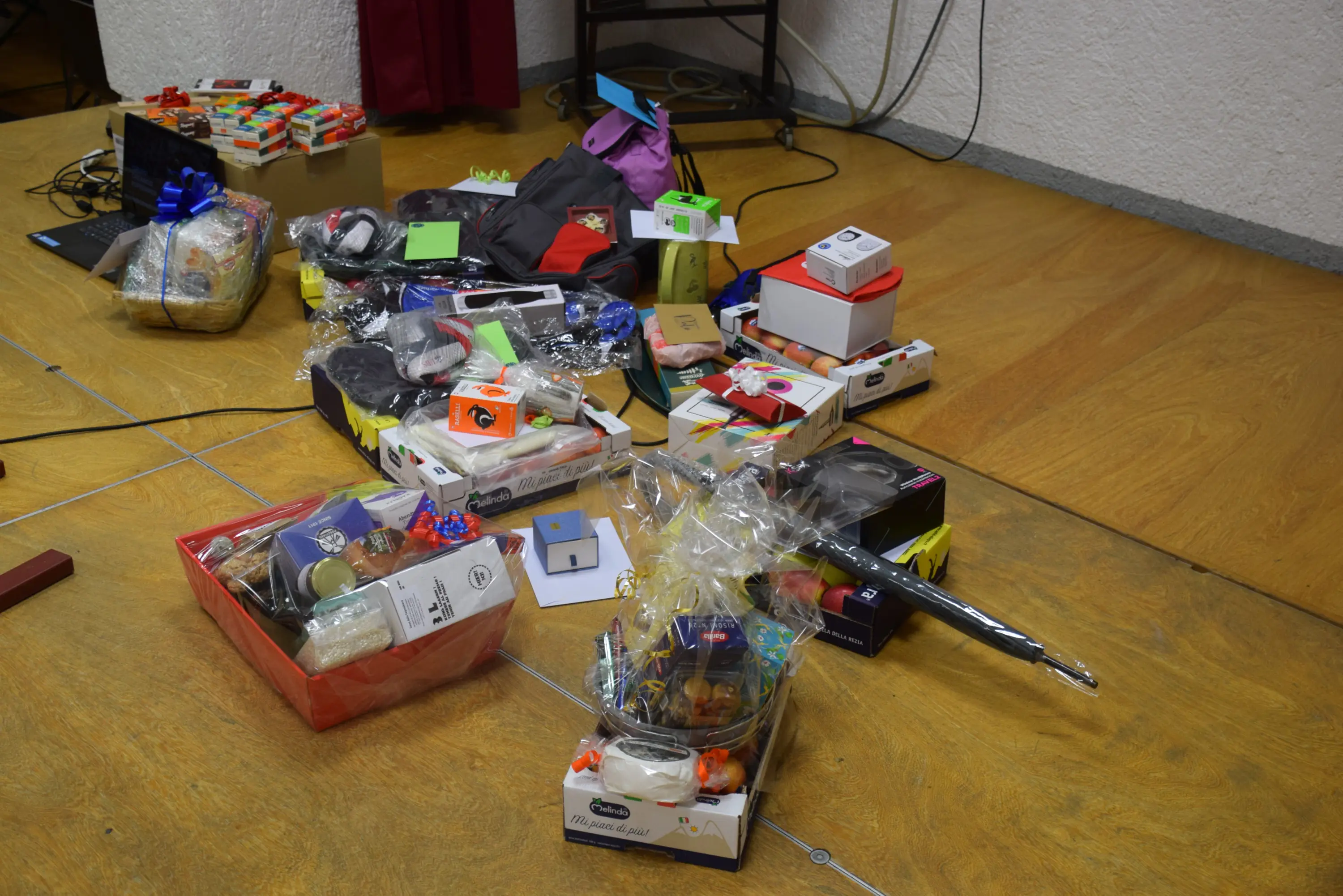 Un assortimento di regali impacchettati e non, disposti su un pavimento in legno. Ci sono scatole colorate, sacchetti e vari oggetti, tra cui cibo e accessori.