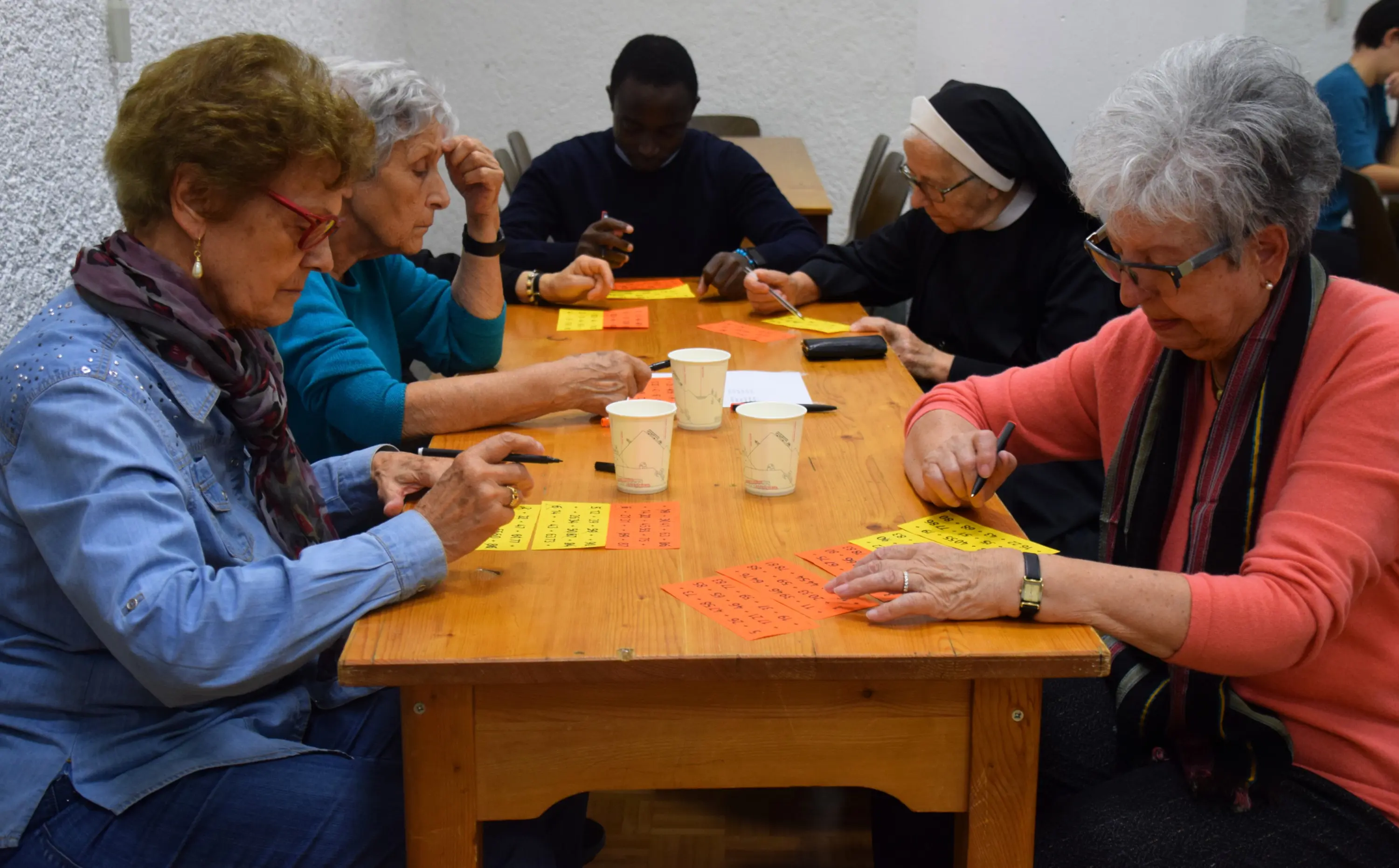 Un gruppo di sei persone, tra cui donne anziane e un uomo, è seduto attorno a un tavolo di legno. Stanno scrivendo su fogli di carta colorati, alcuni gialli e altri arancioni. Sono concentrati e sembrano partecipare a un'attività di gruppo. Sul tavolo ci sono anche bicchieri di carta.