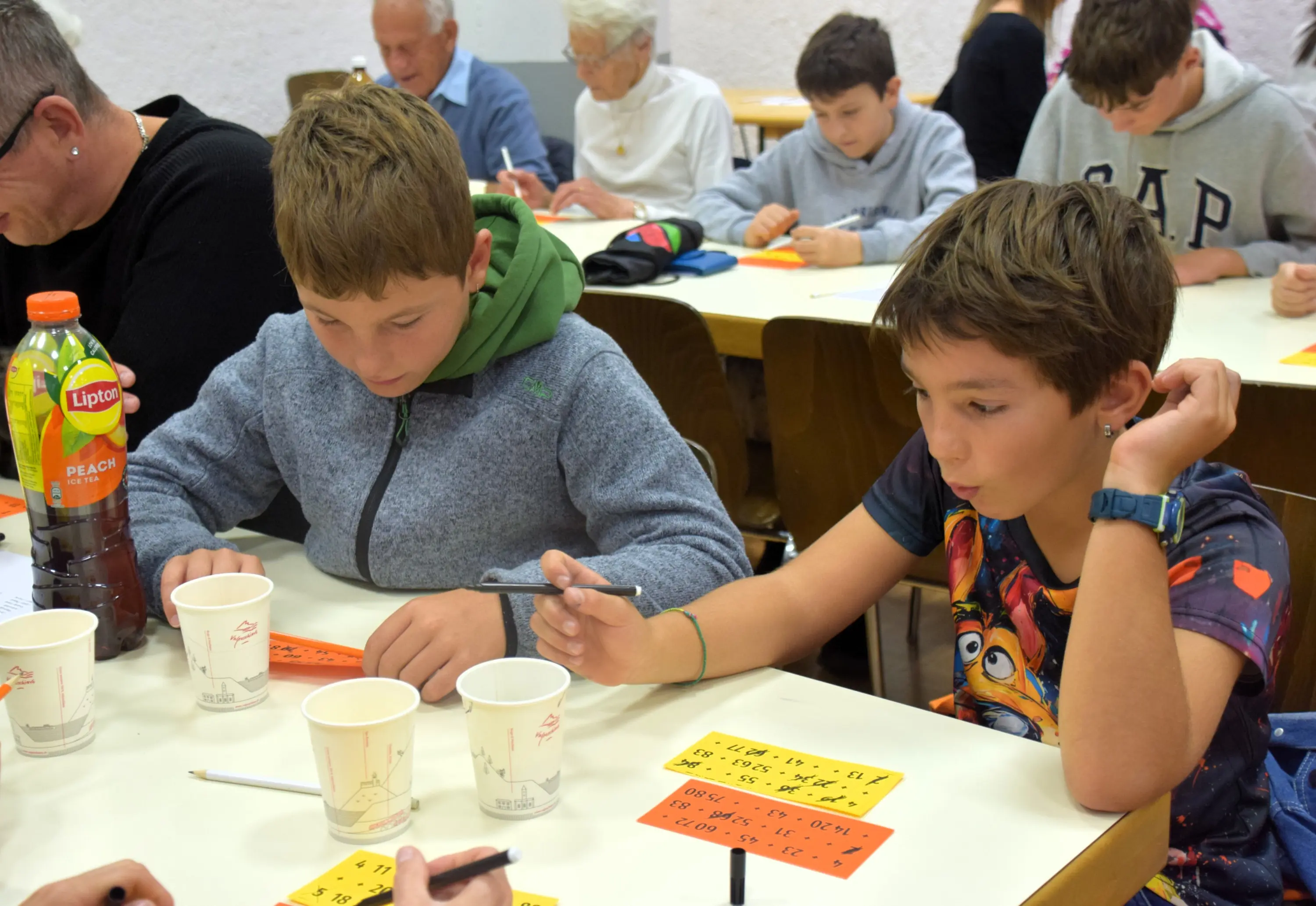 Due ragazzi seduti a un tavolo, concentrati su un gioco. Insieme a loro ci sono adulti e altri bambini. Alcuni partecipanti hanno bordo di carta giallo davanti e tengono penne, mentre sul tavolo ci sono bicchieri di plastica e bottiglie di bevande.