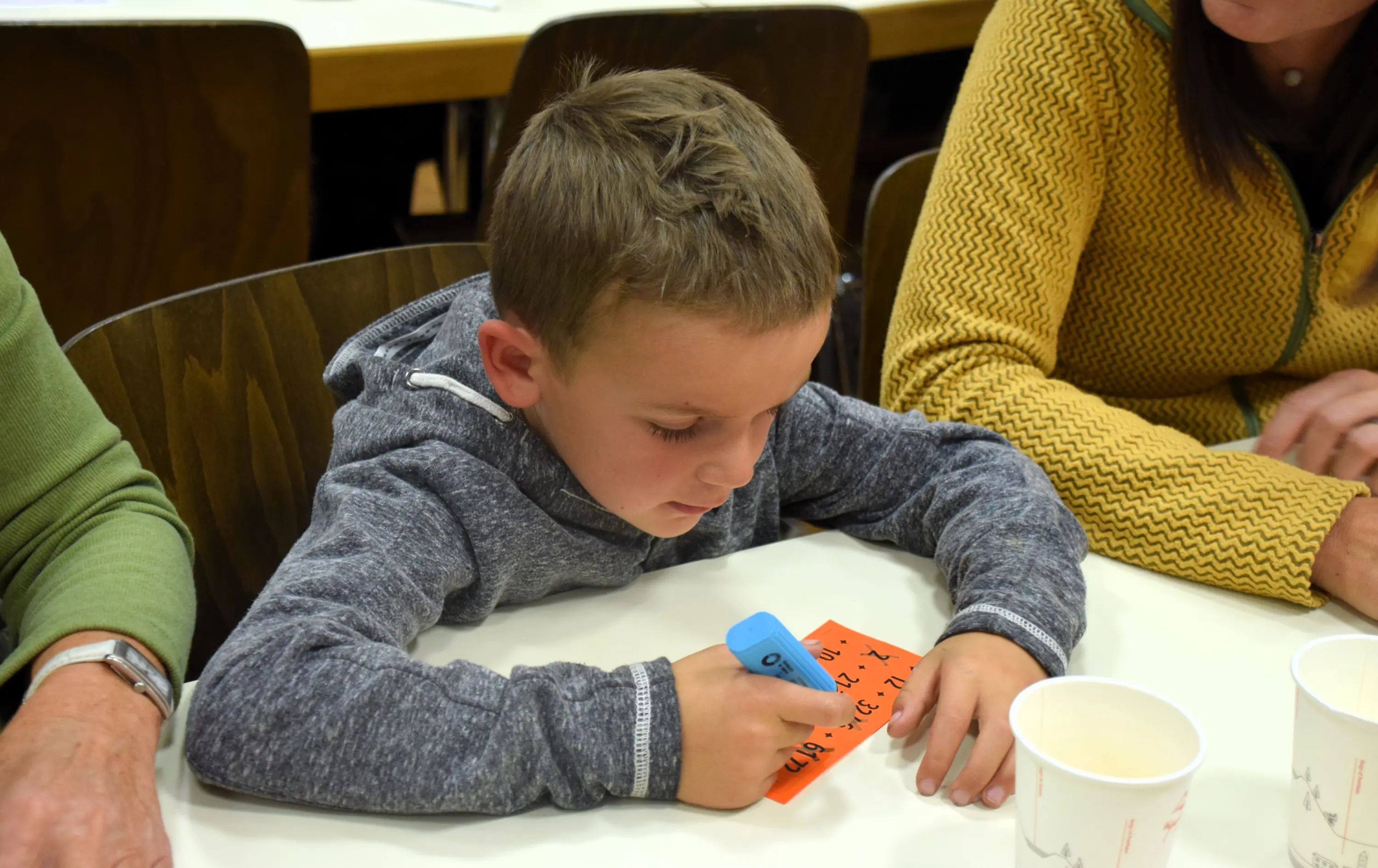 Un bambino seduto a un tavolo scrive su un cartoncino arancione usando una penna blu. Attorno a lui ci sono adulti e bicchieri di carta.