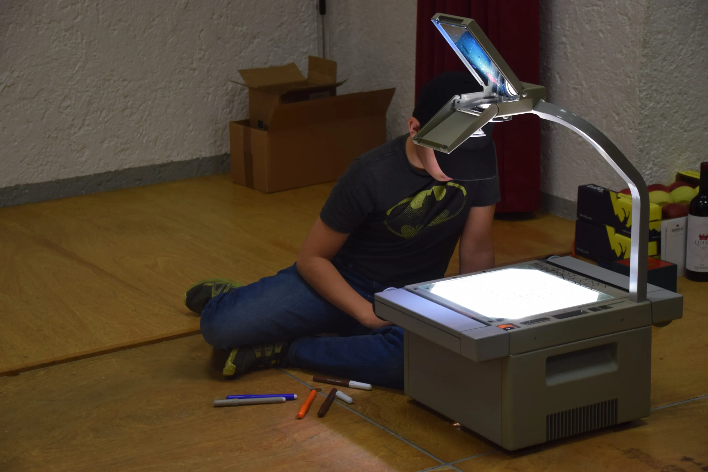 Un ragazzo seduto sul pavimento mentre usa un proiettore per visualizzare dei disegni. Indossa una maglietta con il logo di Batman e ha vari pennarelli colorati accanto a lui.