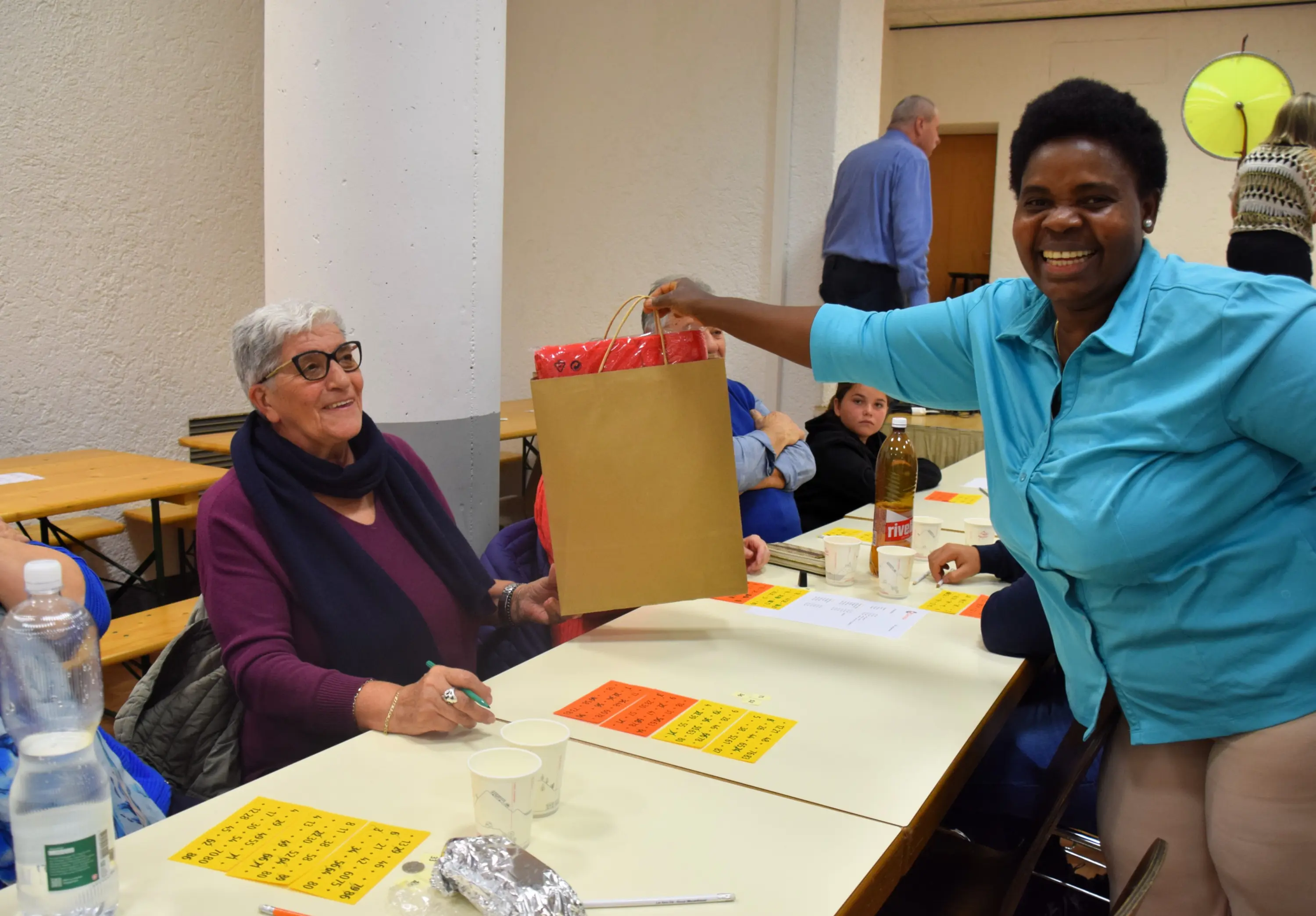 Una donna sorridente porge un sacchetto regalo a un'altra donna seduta a un tavolo. Sul tavolo ci sono alcuni bicchieri e bottiglie, mentre altre persone sono visibili sullo sfondo.