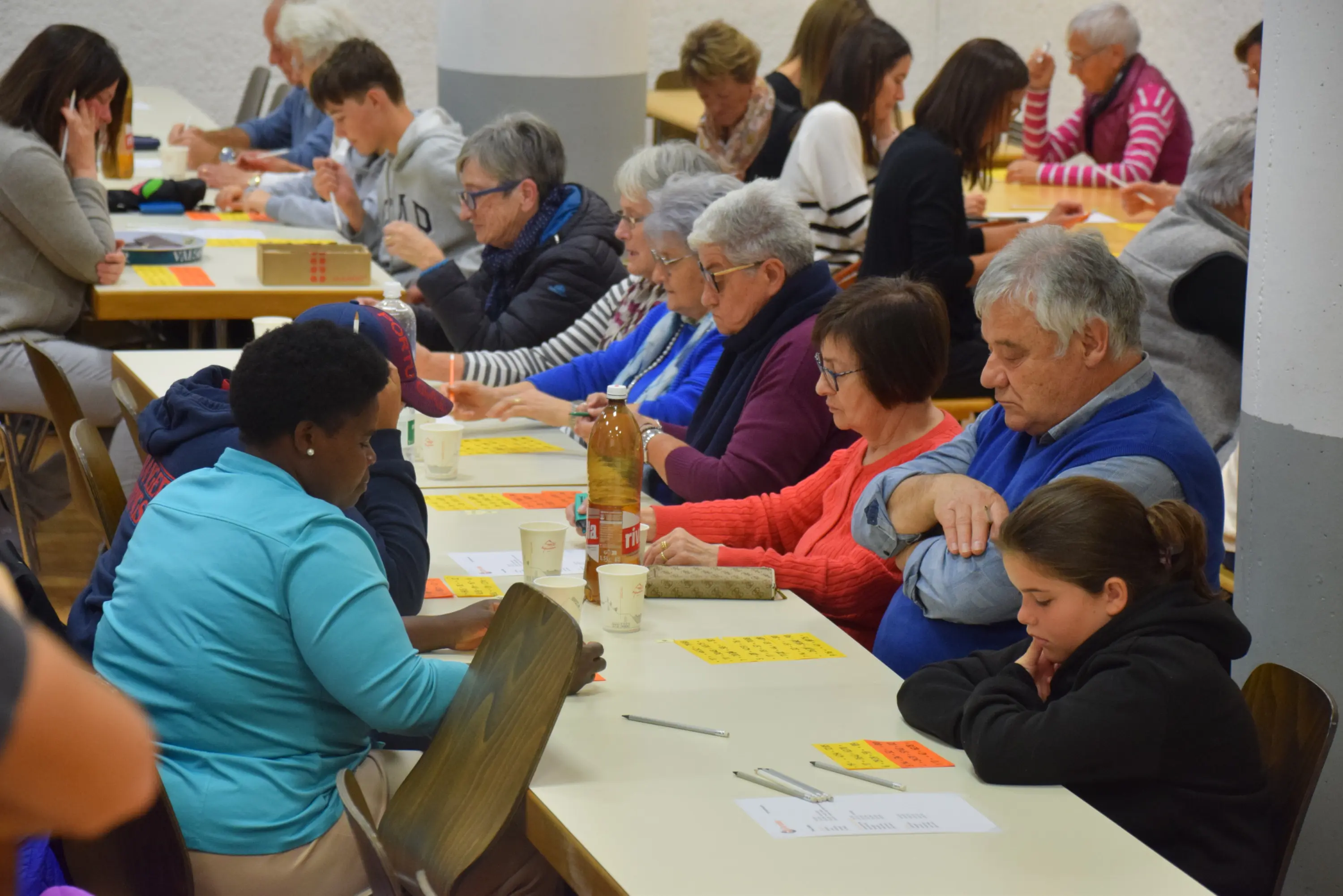 Un gruppo di persone di diverse età, sedute a tavoli, sono concentrati su un gioco. Alcuni hanno cartelle di bingo davanti a loro, mentre altri scrivono o osservano. Sulla tavola ci sono bottiglie e bicchieri, creando un'atmosfera sociale e ricreativa.