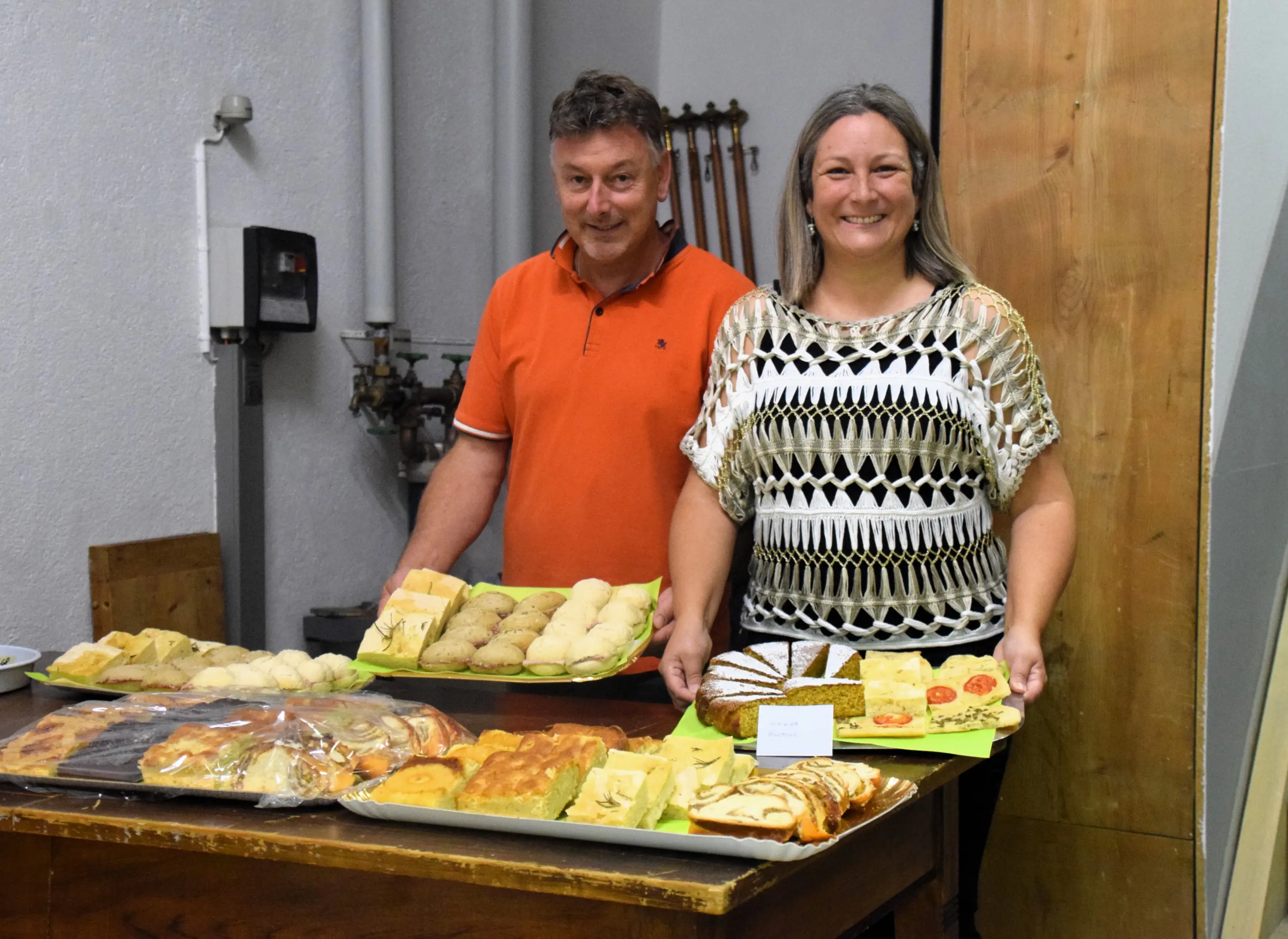 Due persone sorridenti dietro un bancone con una varietà di dolci e prodotti da forno disposti su piatti colorati.