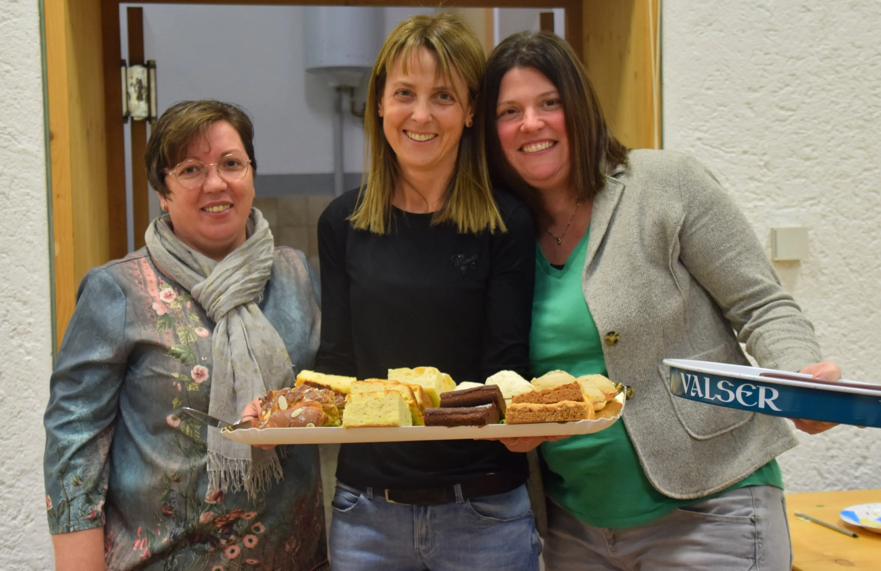 Tre donne sorridenti posano insieme, ognuna tenendo un vassoio con vari dolci e snack. Sullo sfondo, una parete bianca e una porta aperta.