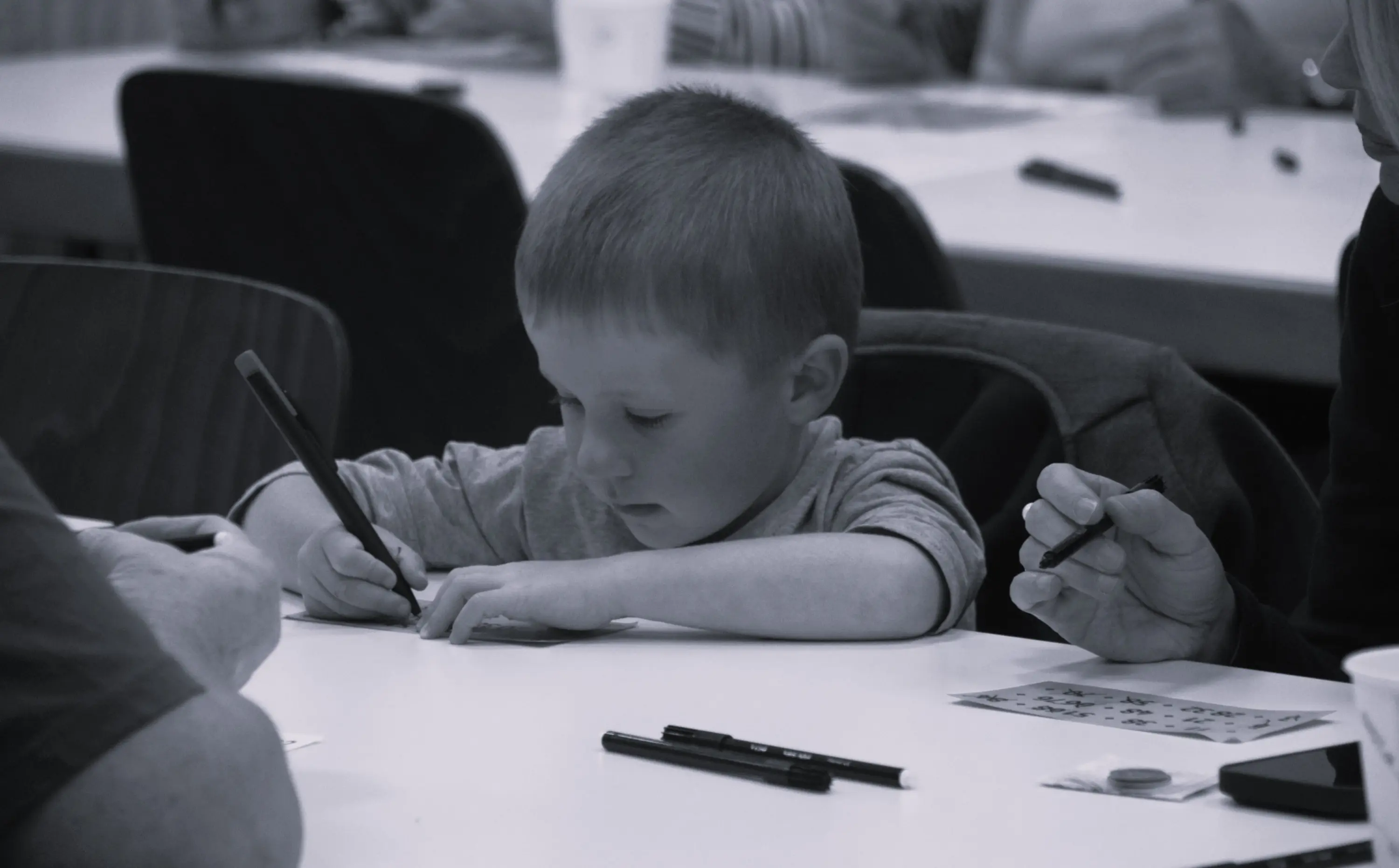 Un bambino sta disegnando su un tavolo, concentrato su un foglio. Si vedono delle penne nere e altre persone intorno che partecipano all'attività.