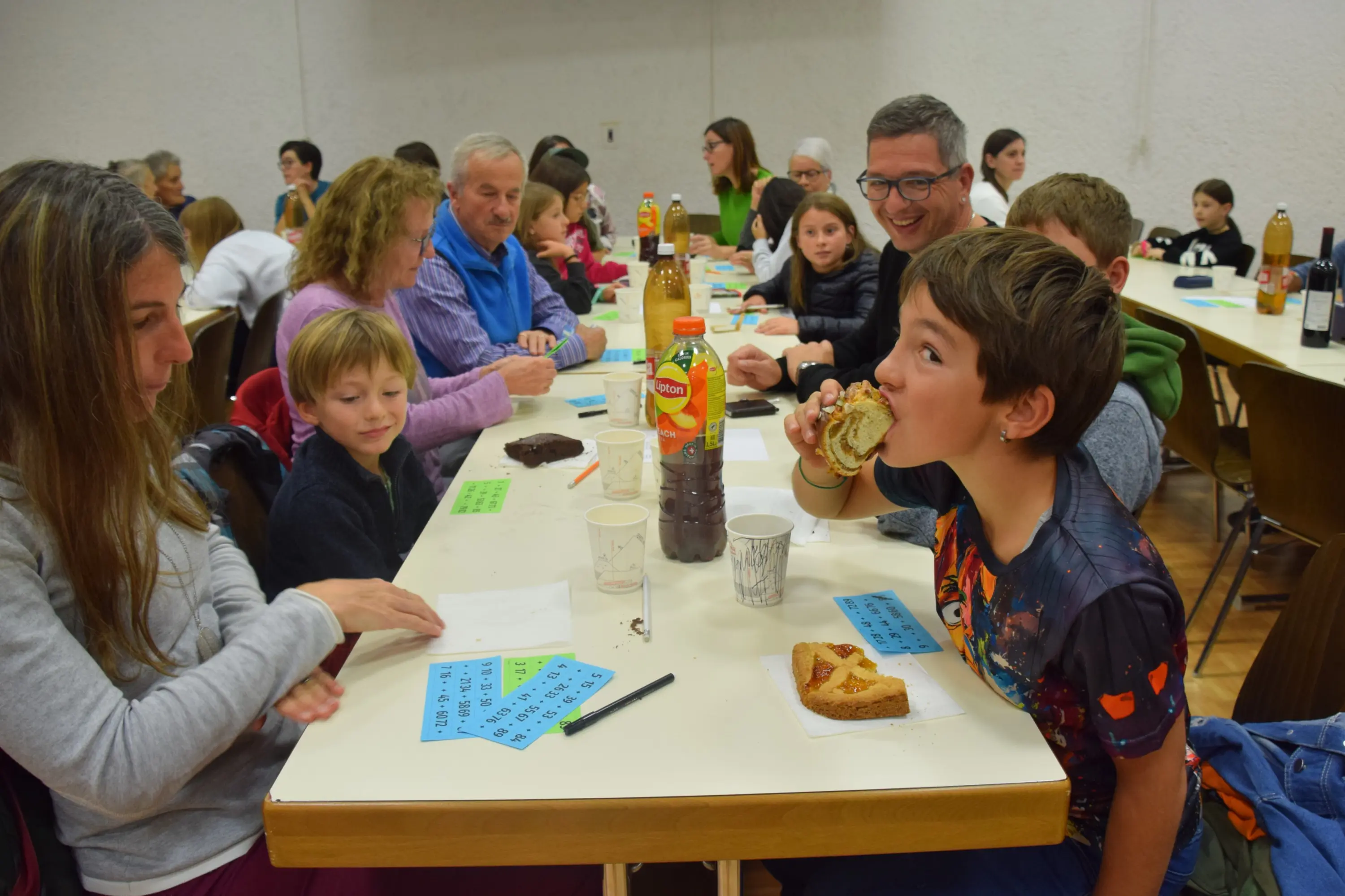 Una sala piena di persone che partecipano a un evento sociale. Alcuni adulti e bambini siedono a lungo tavoli, mentre un ragazzo mangia un panino. Altri partecipanti chiacchierano e ci sono bevande sui tavoli.