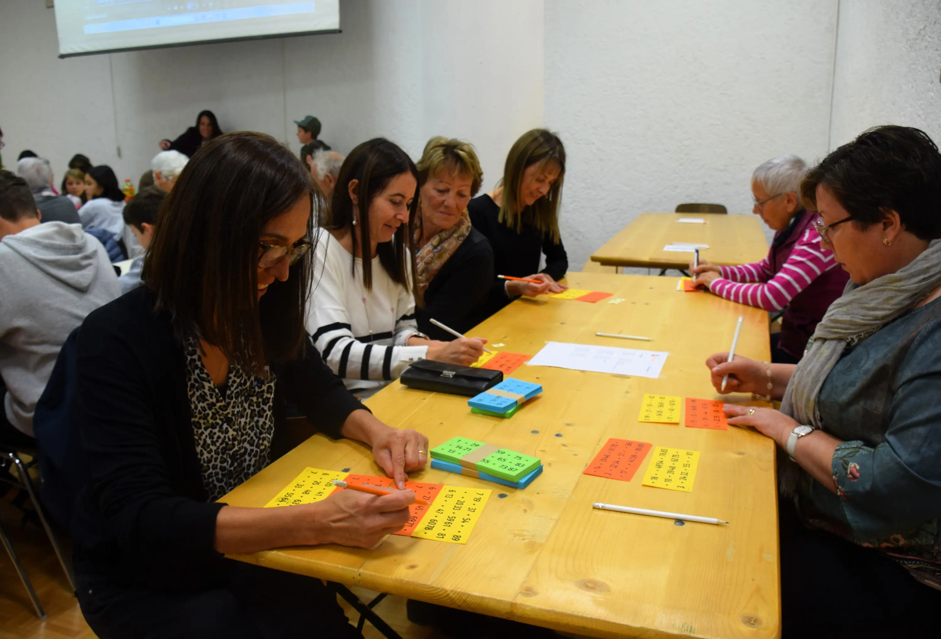 Un gruppo di persone sedute a un tavolo, impegnate a scrivere su fogli colorati e a usare penne. Alcuni partecipanti sorridono e si concentrano sulle loro attività, mentre altri guardano i loro materiali. Sullo sfondo si intravede un proiettore acceso.