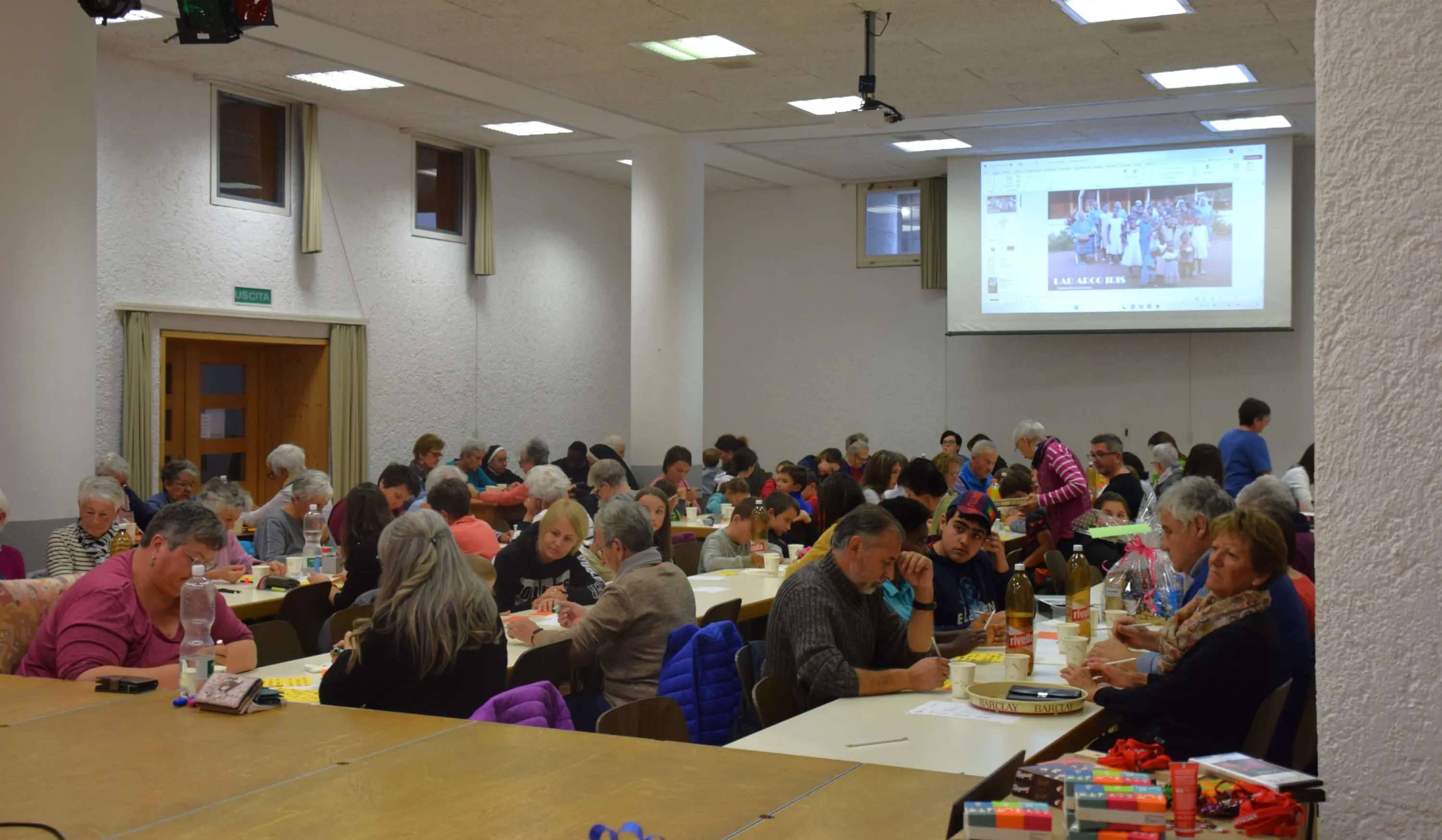 Sala affollata con molte persone sedute a tavoli. Alcuni partecipanti sono intenti a scrivere o parlare, mentre un grande schermo proietta immagini sul fondo. L'atmosfera sembra conviviale e animata.