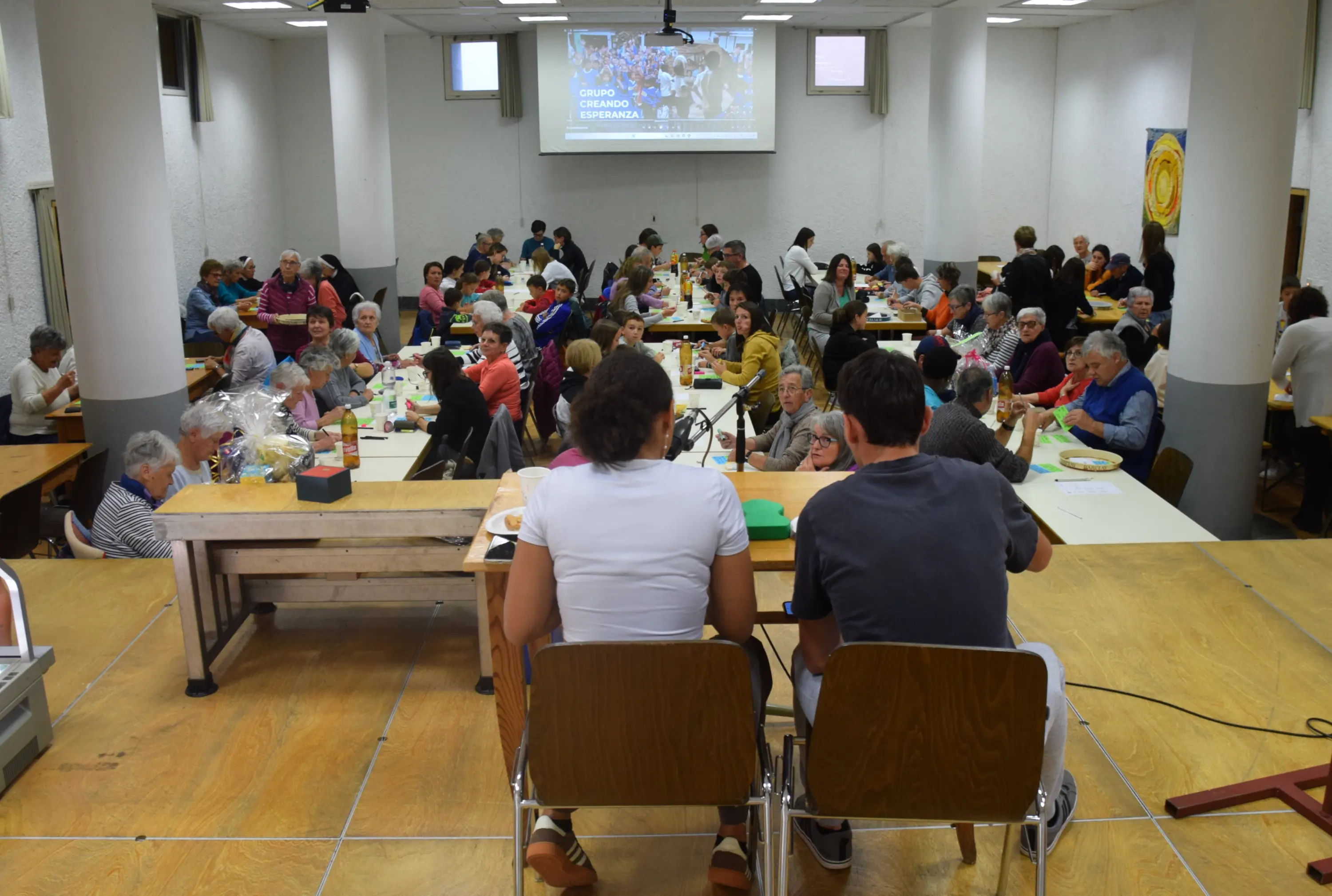 Una sala affollata con persone sedute a lungo tavoli mentre partecipano a un evento. Due giovani seduti in primo piano guardano verso il pubblico. Sullo sfondo, un muro con uno schermo e altre persone si divertono.
