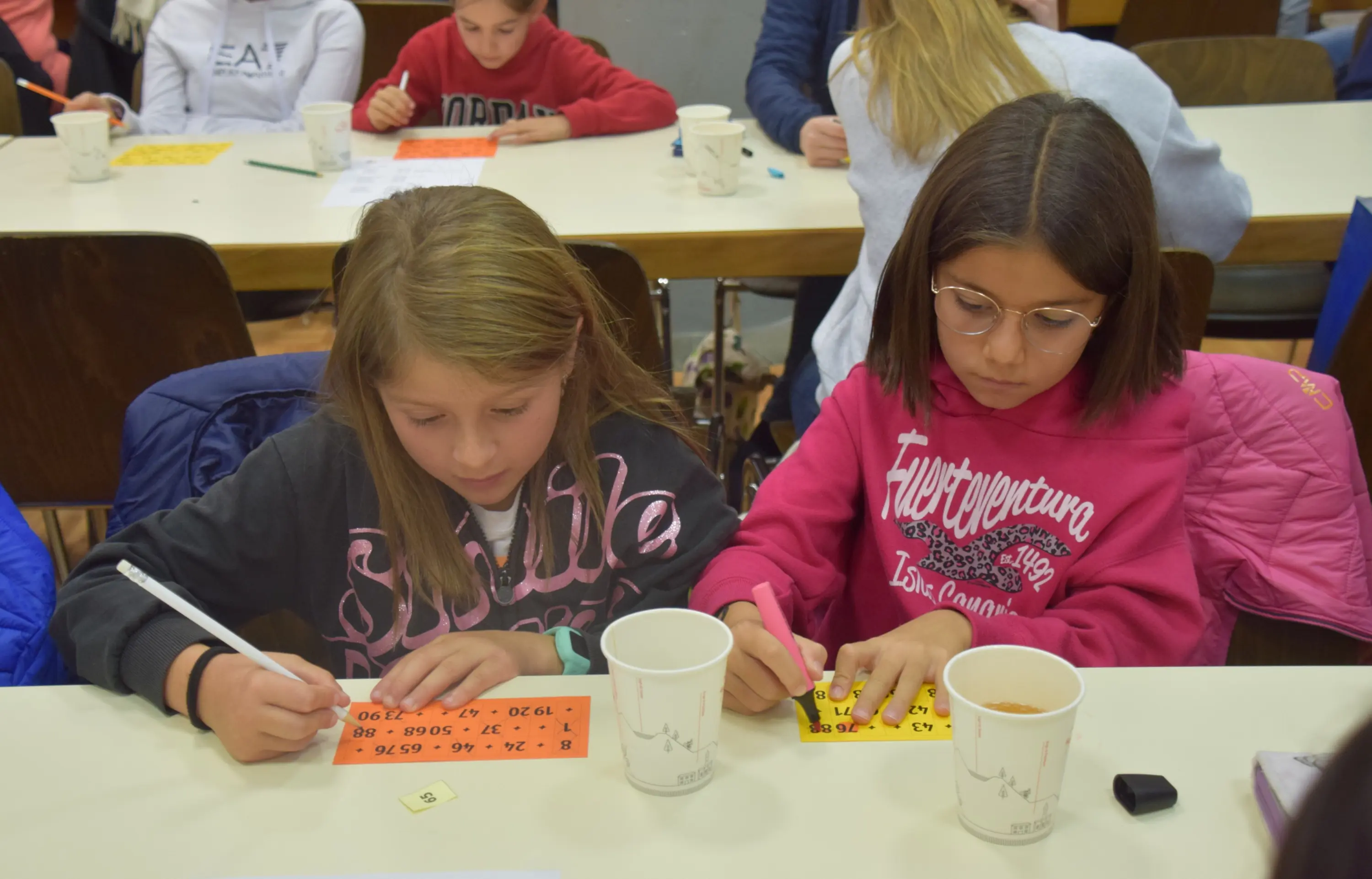 Due ragazze sedute a un tavolo, una scrive su un cartellone arancione mentre l'altra segna su una scheda di gioco, entrambe sono concentrare. Sulla tavola ci sono tazze di carta.