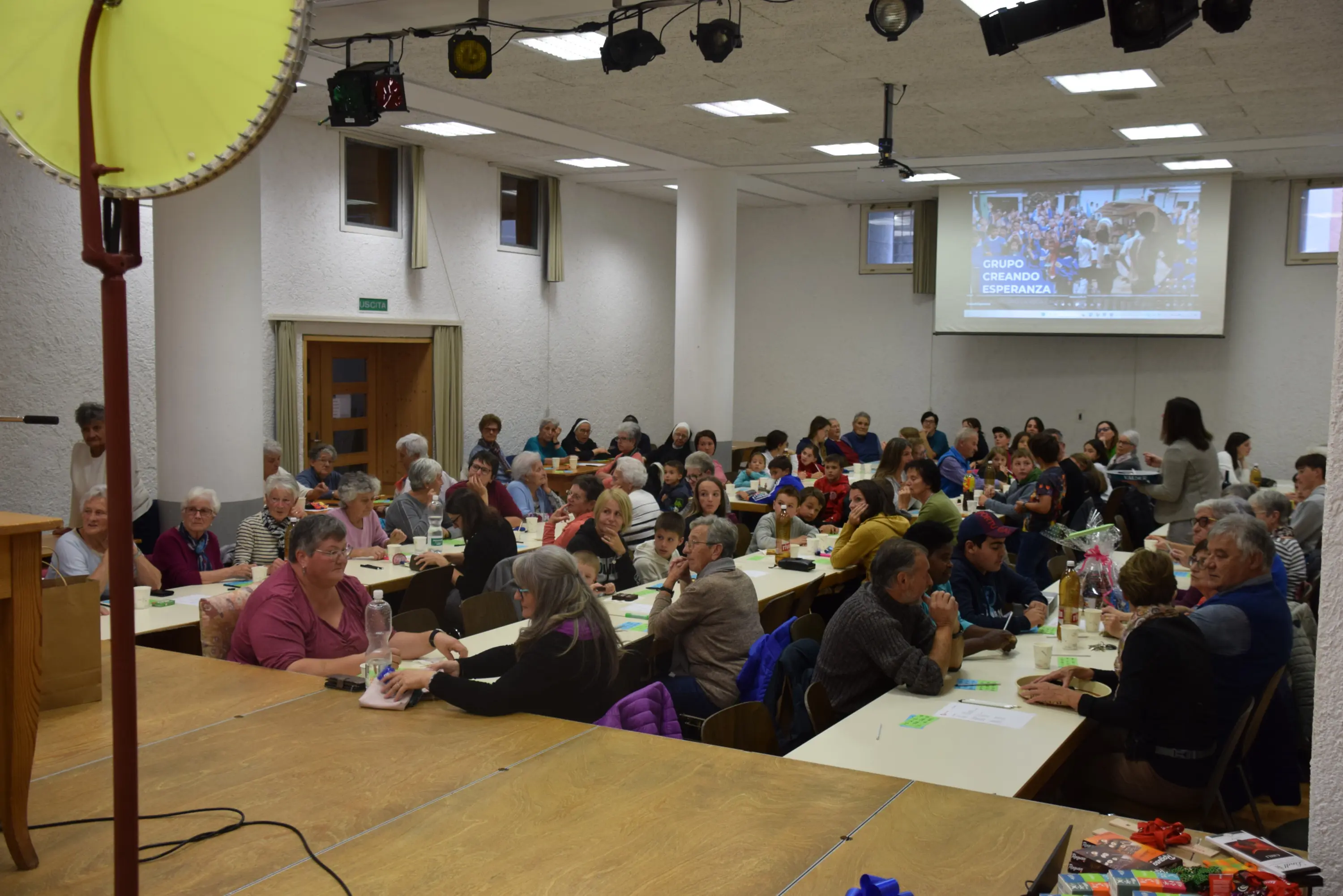 Una sala affollata con persone sedute a tavoli, partecipando a un evento. Alcuni stanno interagendo tra loro, mentre un proiettore mostra un'immagine sullo sfondo. L'atmosfera è vivace e conviviale.