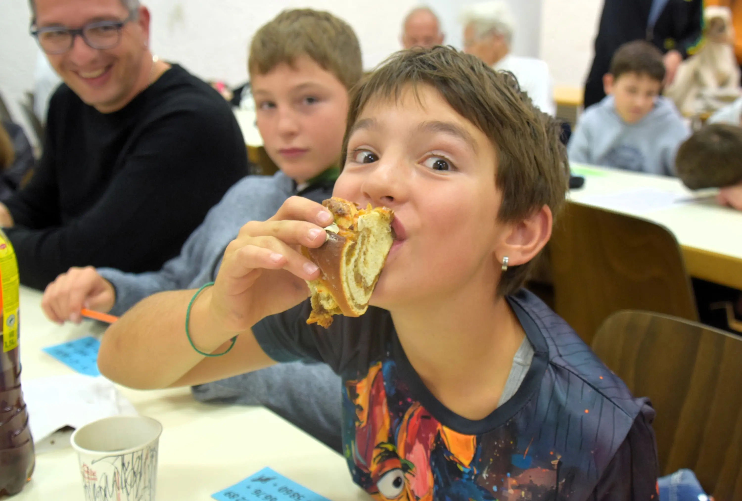 Un ragazzo sorride mentre mangia una fetta di torta, con una mano sulla bocca. Sullo sfondo si vedono altre persone che stanno partecipando a un evento.