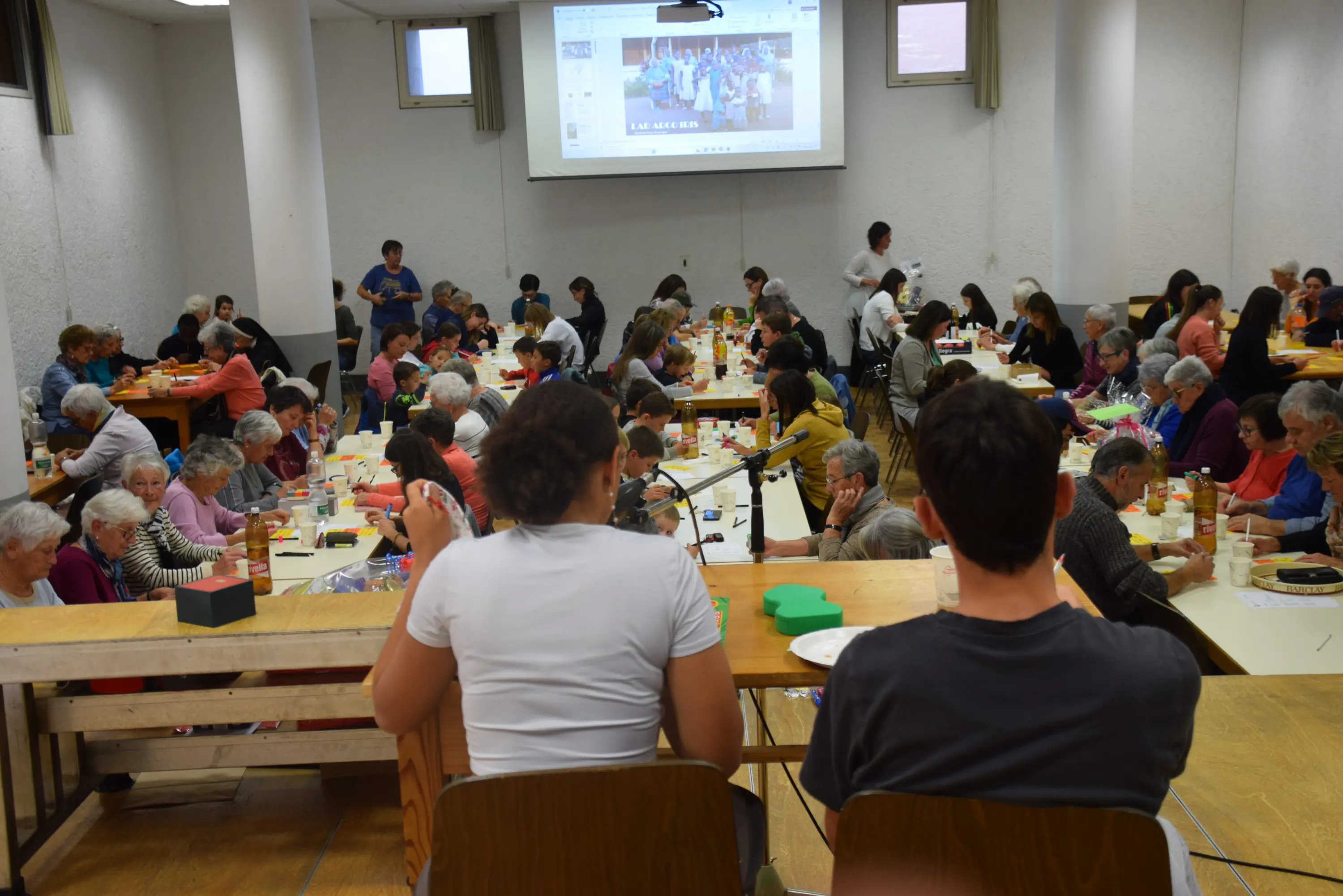 Aula affollata con molte persone anziane sedute a tavoli, mentre un gruppo di giovani sta sul palco. Sullo sfondo, una proiezione video mostra un evento. Atmosfera sociale e conviviale.