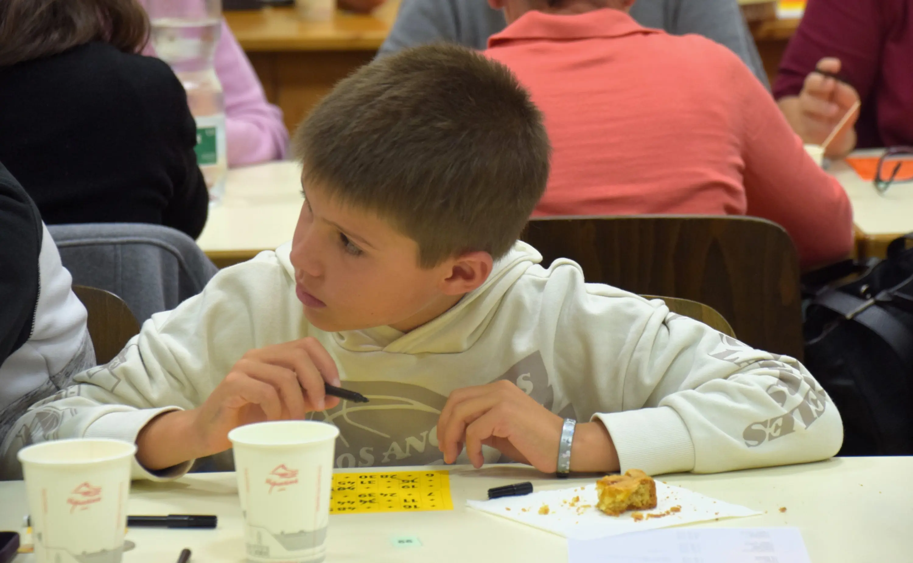 Un ragazzo con una felpa chiara, intento a scrivere su un foglio mentre mangia un biscotto. Altri partecipanti, sfocati sullo sfondo, sono seduti a un tavolo.