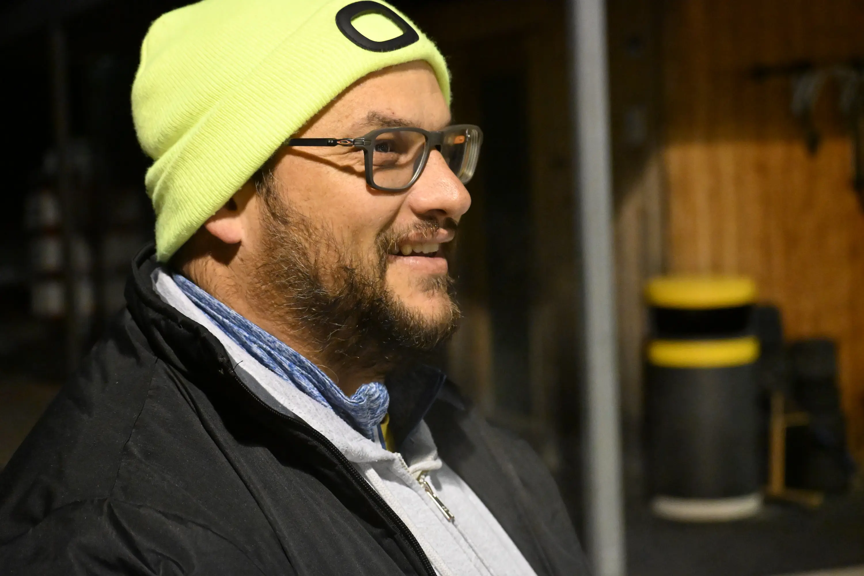 Uomo con occhiali e cappello giallo fluo, sorridente, indossando un giubbotto scuro, in un ambiente esterno. Sullo sfondo si