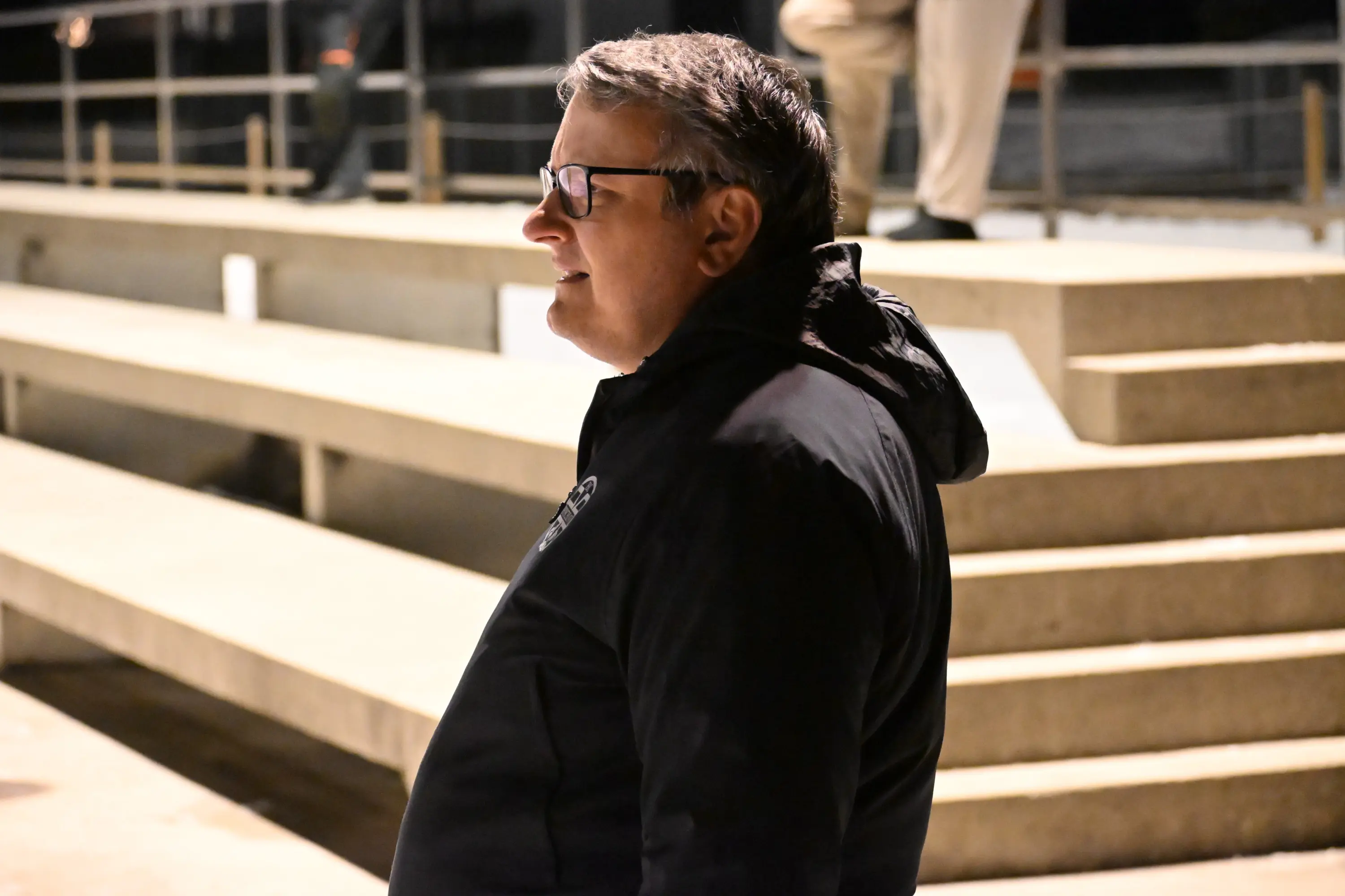 Una persona in giacca nera osserva l'ambiente circostante, con una postura rilassata. Sullo sfondo si vedono gradini e altri individui.