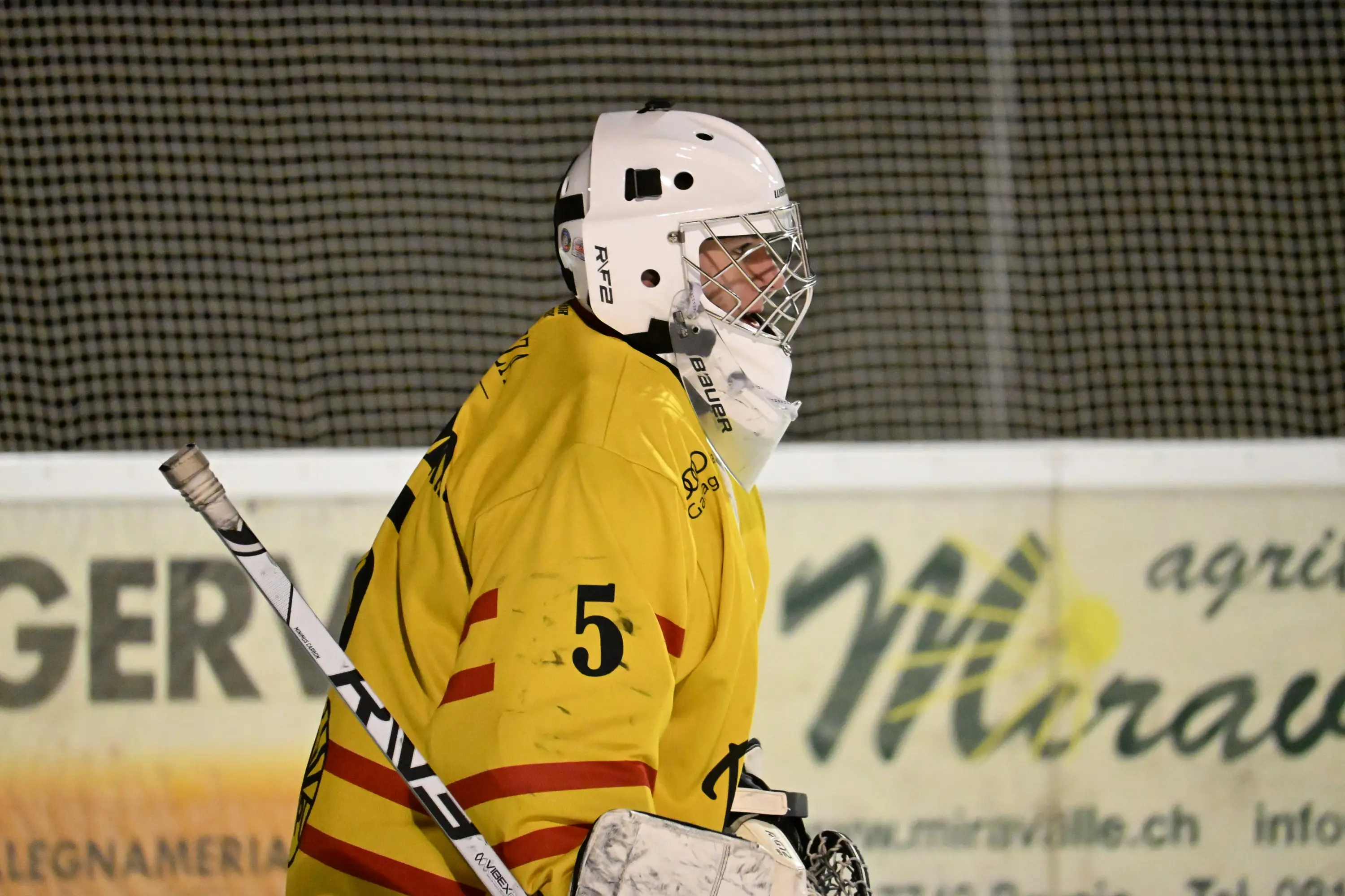 Giocatore di hockey su ghiaccio con un casco bianco e una maglia gialla, che guarda verso il ghiaccio. Sullo sfondo si intravede la rete della porta e