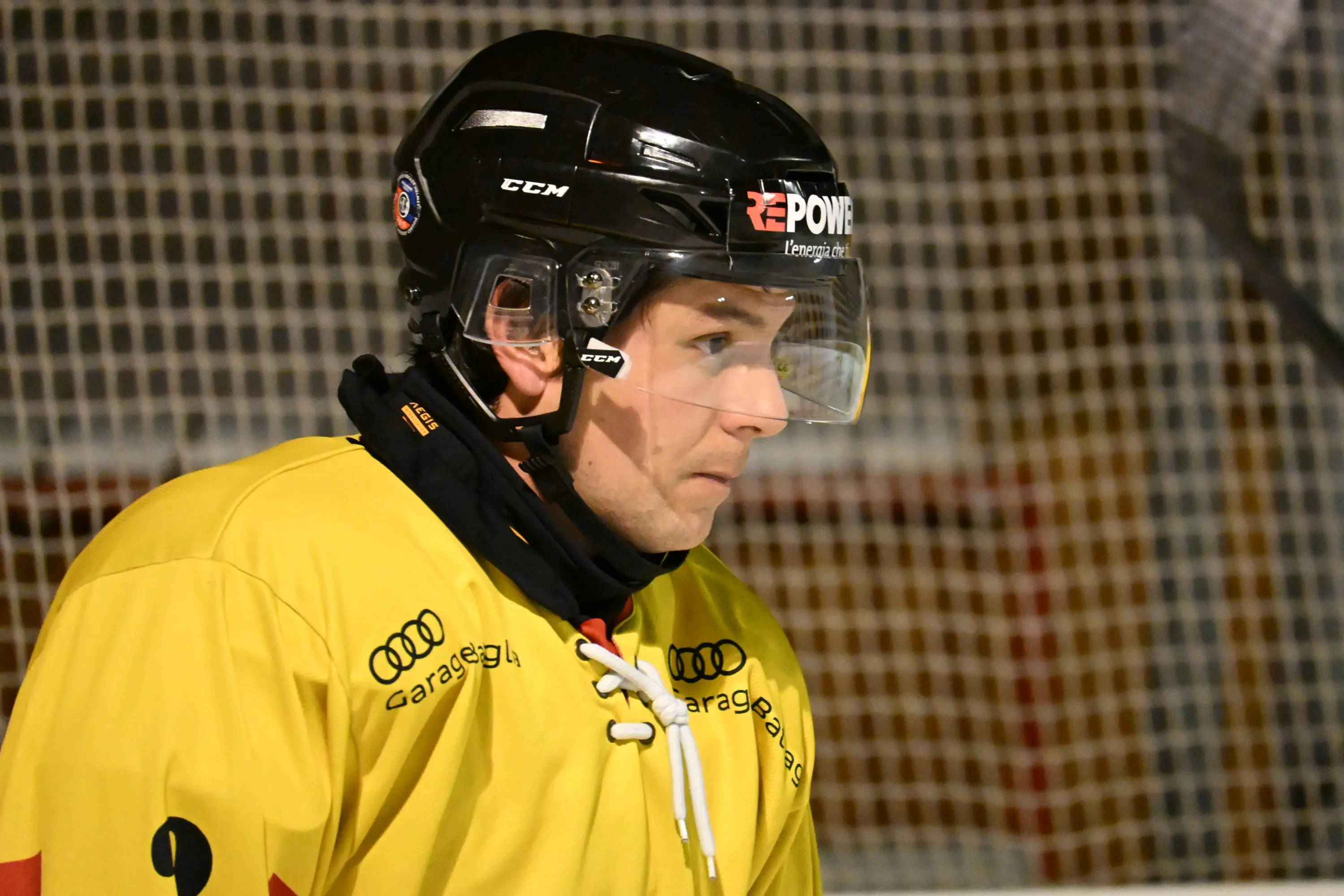 Giocatore di hockey su ghiaccio in maglia gialla e casco protettivo, concentrato e pronto in campo. La rete della porta è visibile sullo sfondo.