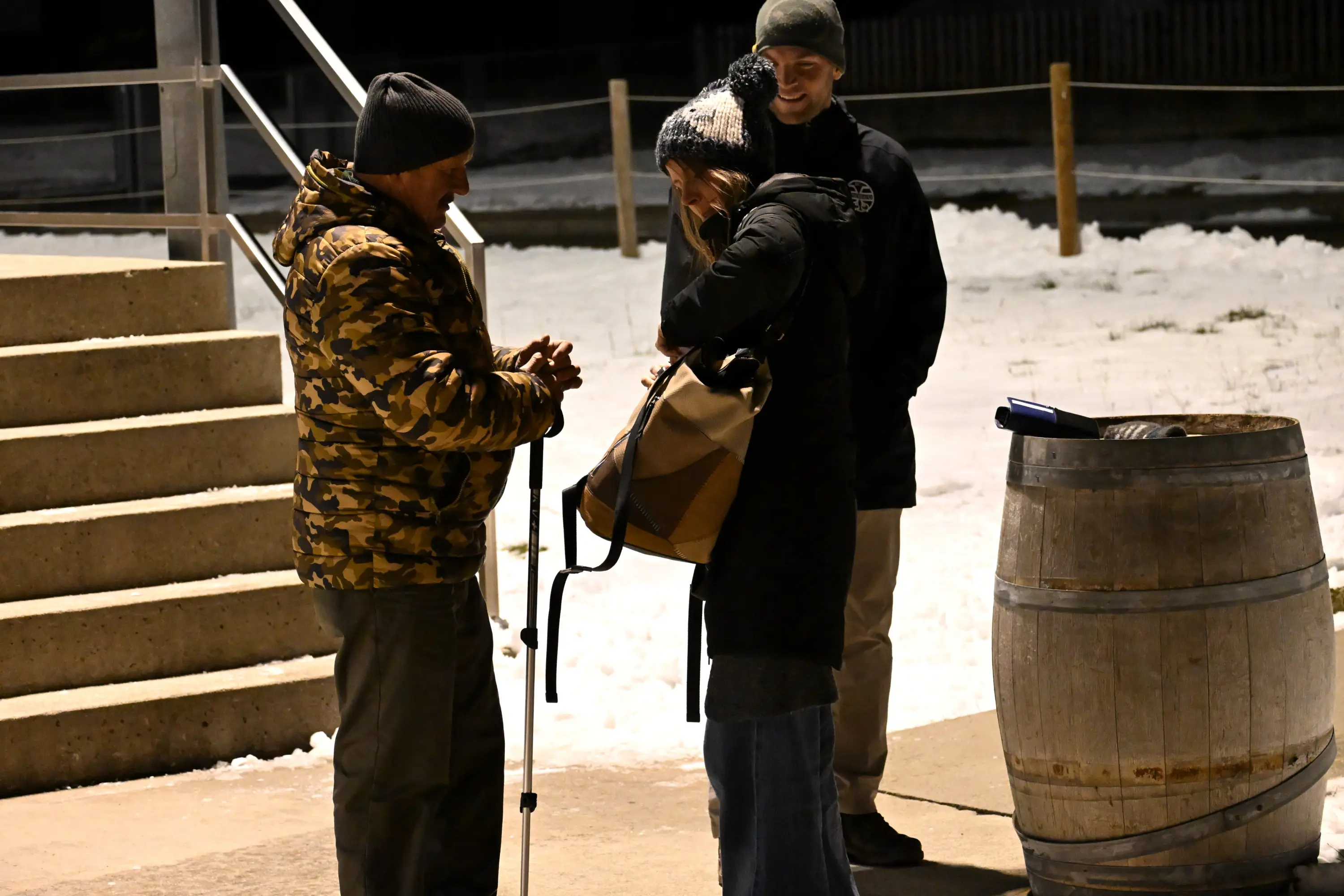 Un uomo con una giacca mimetica e un bastone parla con una donna che tiene una borsa, mentre un altro uomo in giacca nera osserva. Fondo innevato e