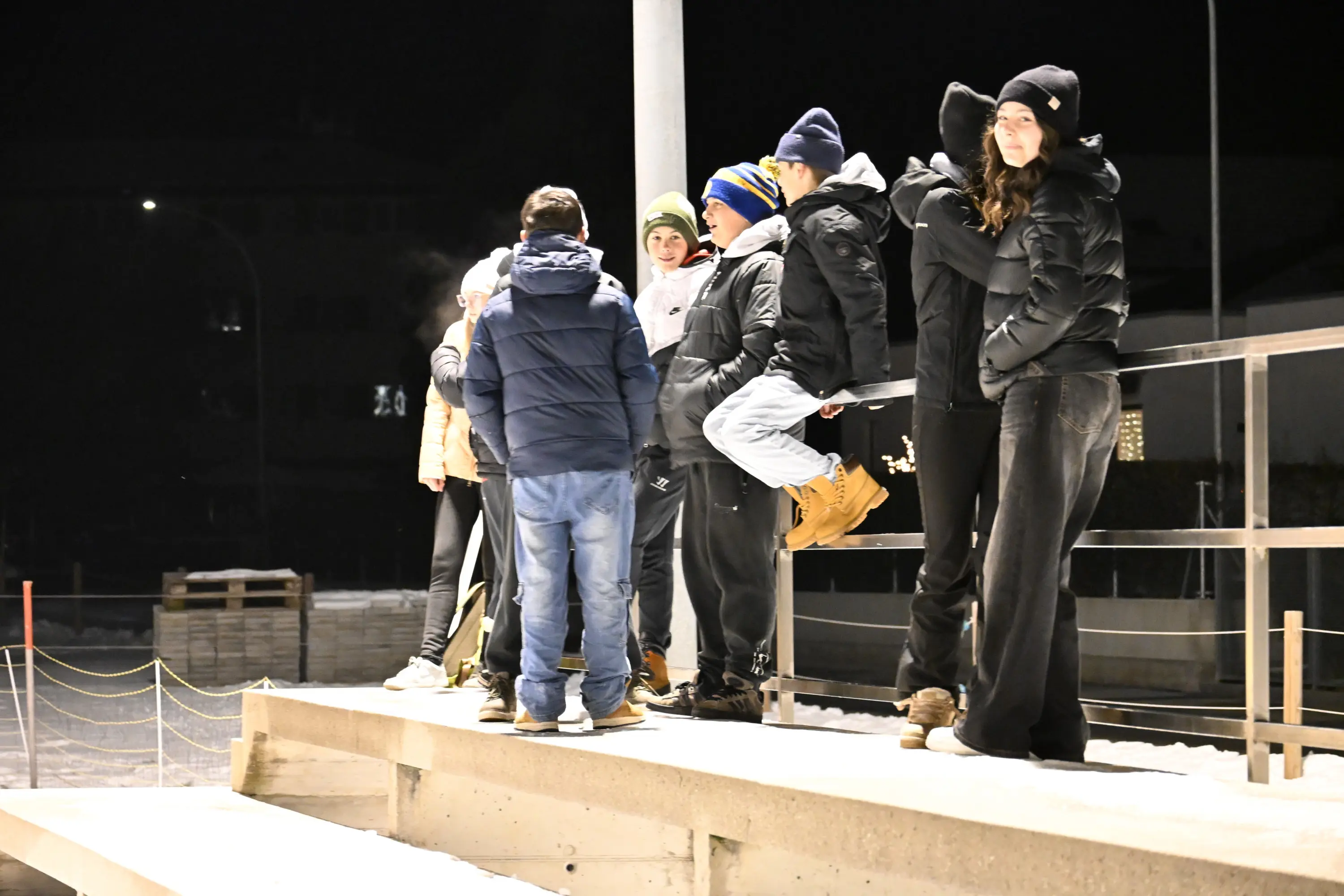 Un gruppo di giovani si riunisce su un muretto all'aperto di notte, indossando giacche e cappelli invernali. Alcuni chiacchierano mentre