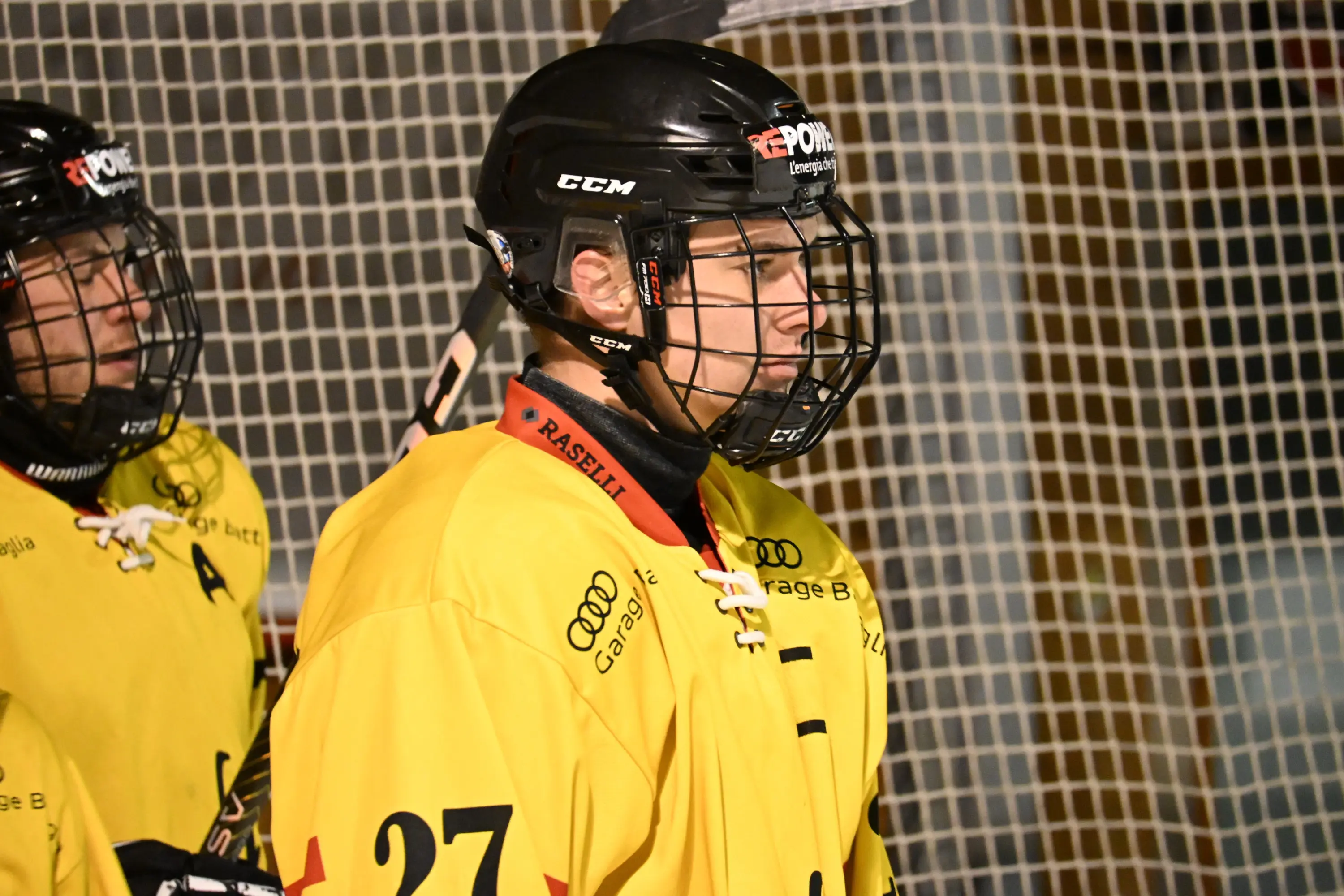 Giocatori di hockey su ghiaccio in divisa gialla, con elmetti neri e grate, si preparano per la partita. Uno dei giocatori indossa il numero 27