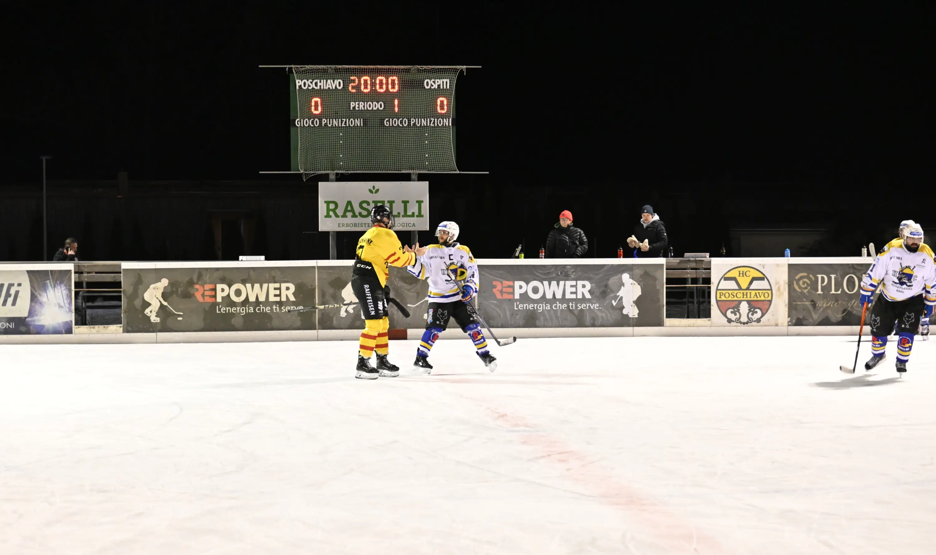 Giocatori di hockey su ghiaccio in azione su un campo illuminato. In primo piano, un giocatore in maglia gialla affronta un avversario, mentre sullo sf