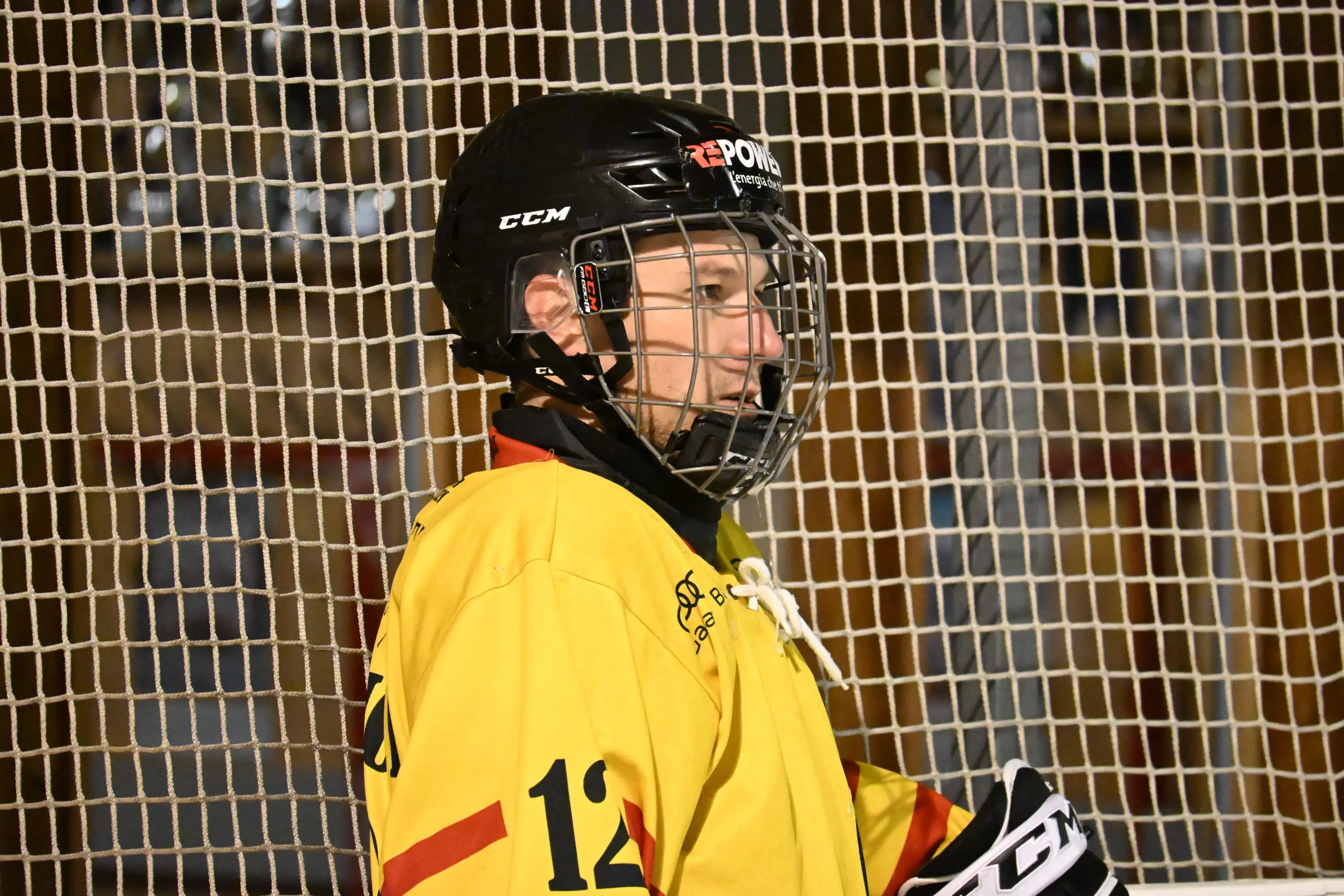 Giocatore di hockey su ghiaccio con casco e maglia gialla, in posa accanto alla rete. Sorriso e concentrazione sul viso.