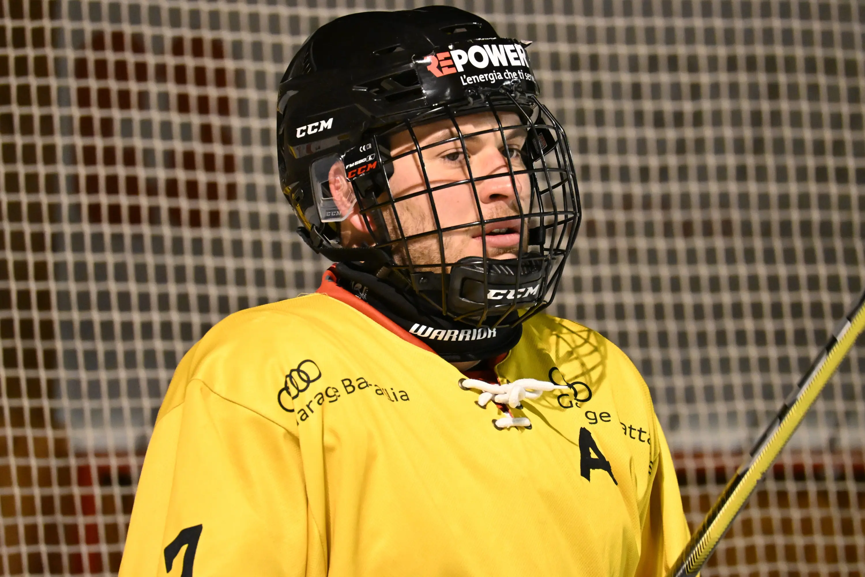 Giocatore di hockey su ghiaccio in divisa gialla, con casco e protezioni, che osserva il campo. Sullo sfondo, la rete della porta.