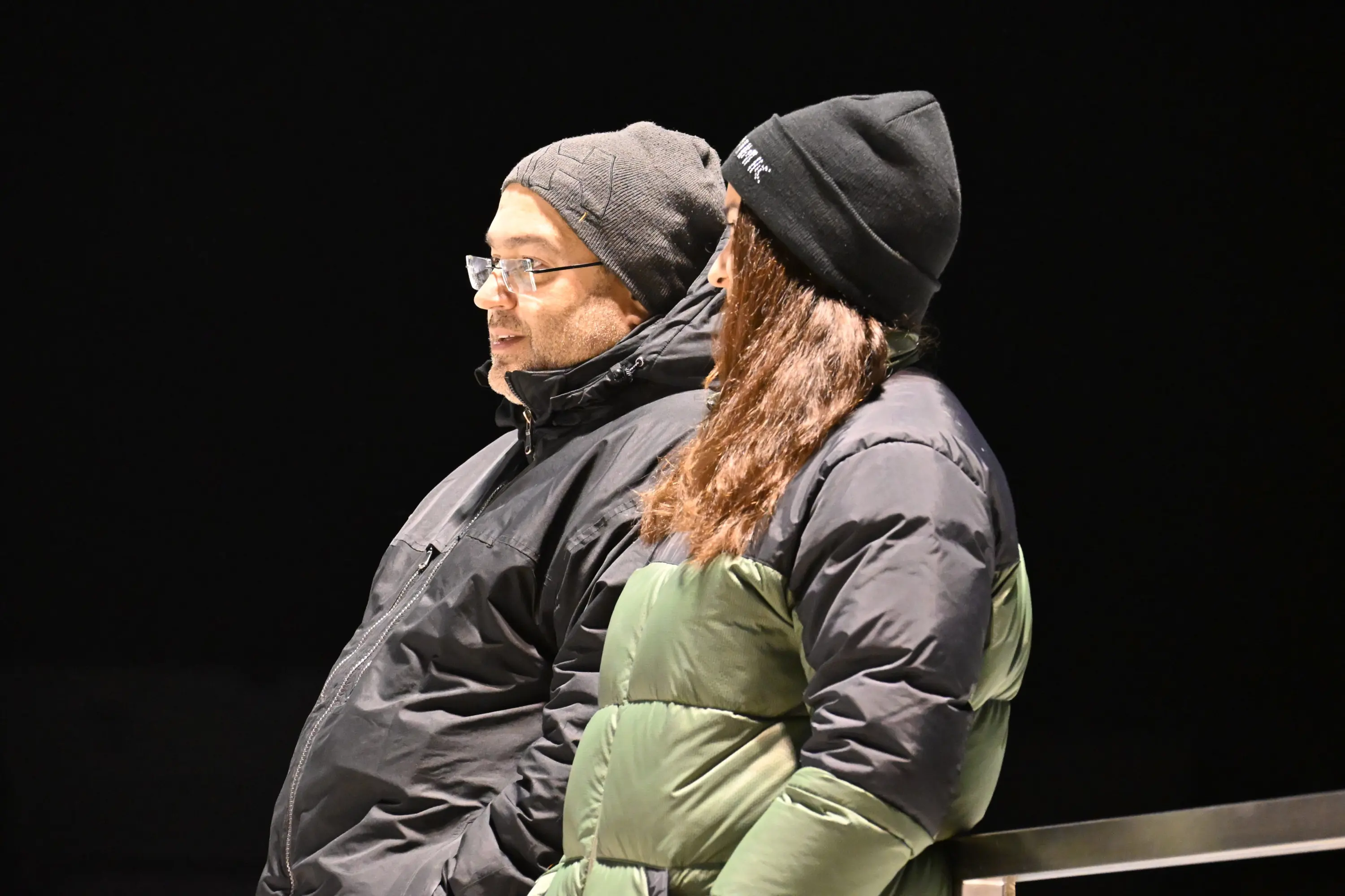 Due persone in abiti invernali, una con occhiali e berretto, l'altra con cappotto imbottito e berretto, osservano l'oscurità della notte.