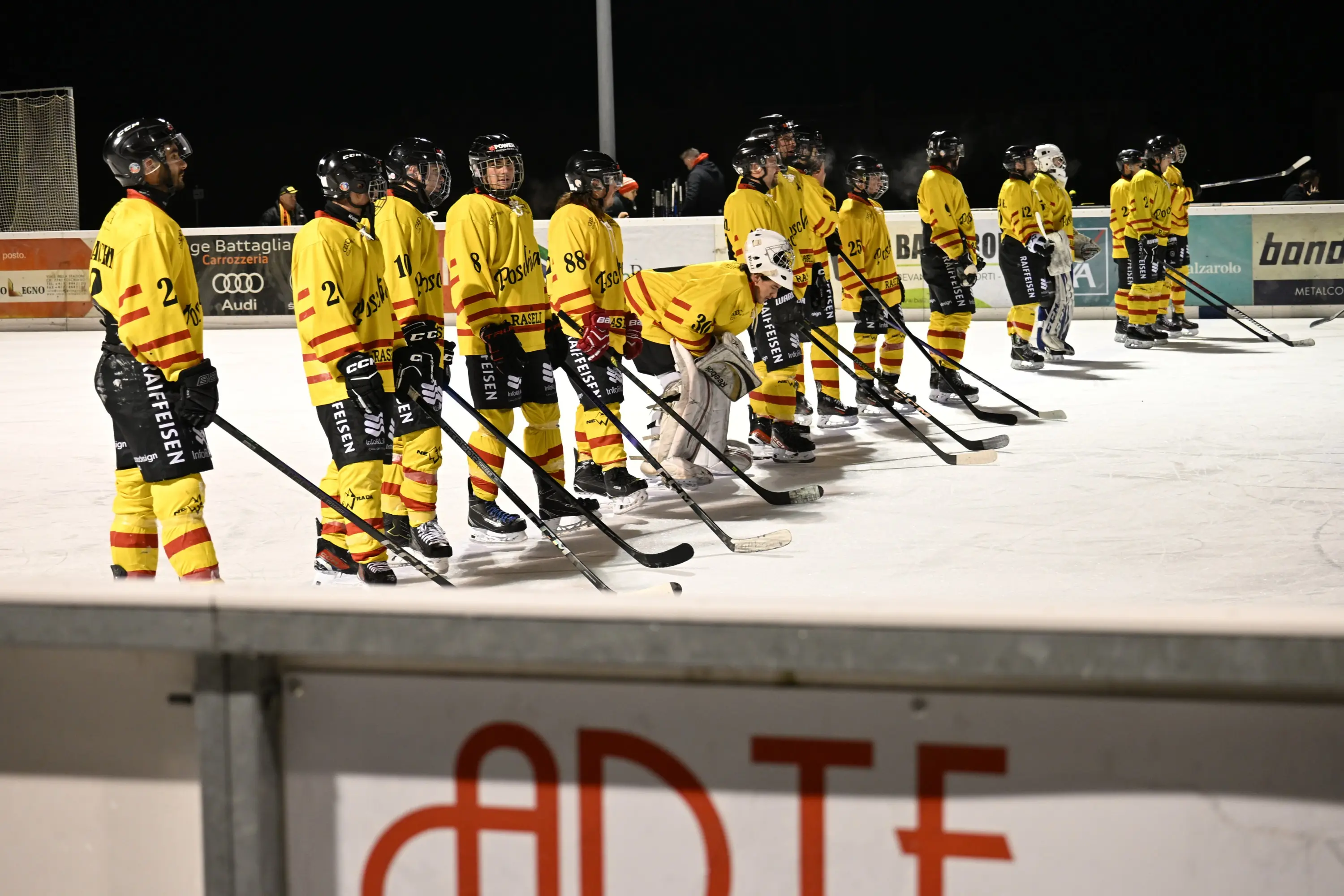 Squadra di hockey su ghiaccio in uniforme gialla e rossa, schierata lungo il bordo della pista. I giocatori indossano caschi e stringono i bastoni da hockey