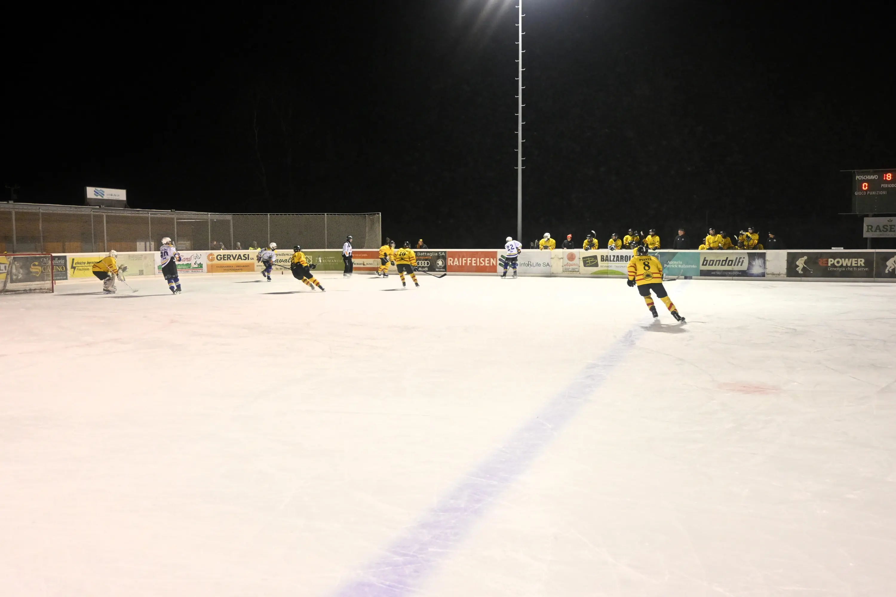 Giocatori di hockey su ghiaccio in azione su un campo illuminato. Alcuni atleti indossano maglie gialle, mentre altri sono in uniforme bianca. In secondo