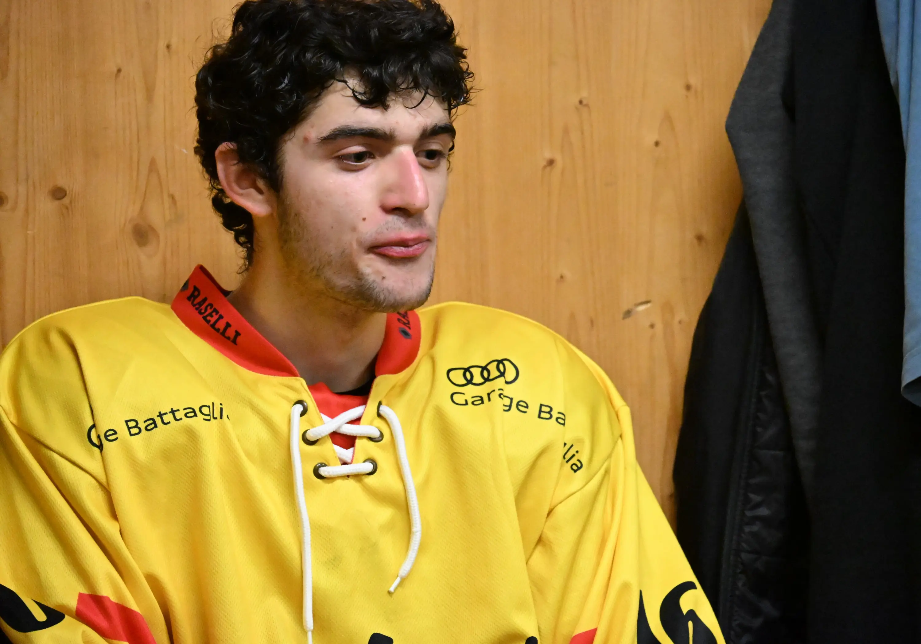 Giovane atleta in una maglia da hockey gialla, seduto in uno spogliatoio con pareti di legno. Ha i capelli scuri e ricci, e sembra pensier