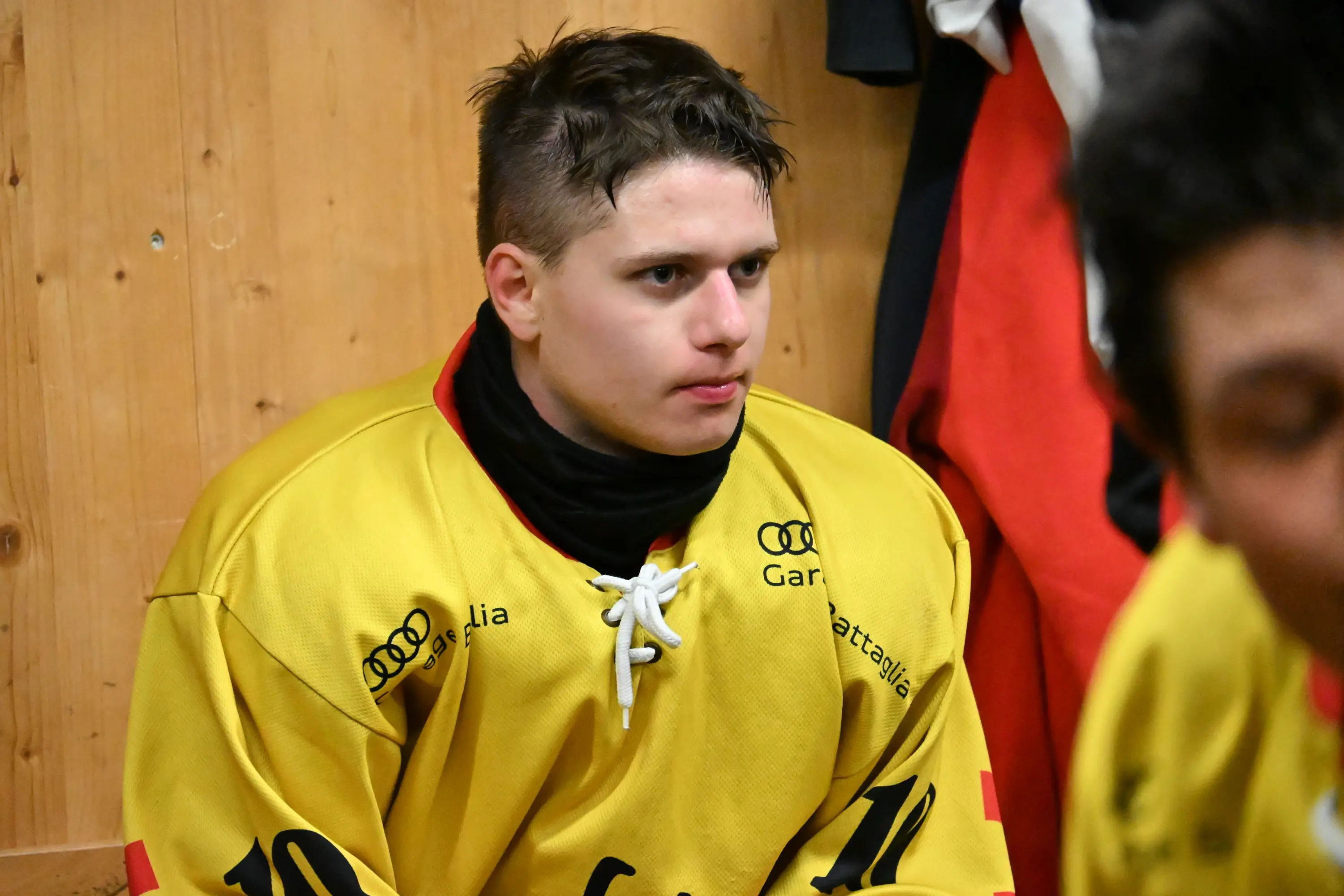 Giocatore di hockey seduto, indossa una maglia gialla con il numero 10. È concentrato e sembra aspettare il proprio turno, con uno sfondo di pareti in