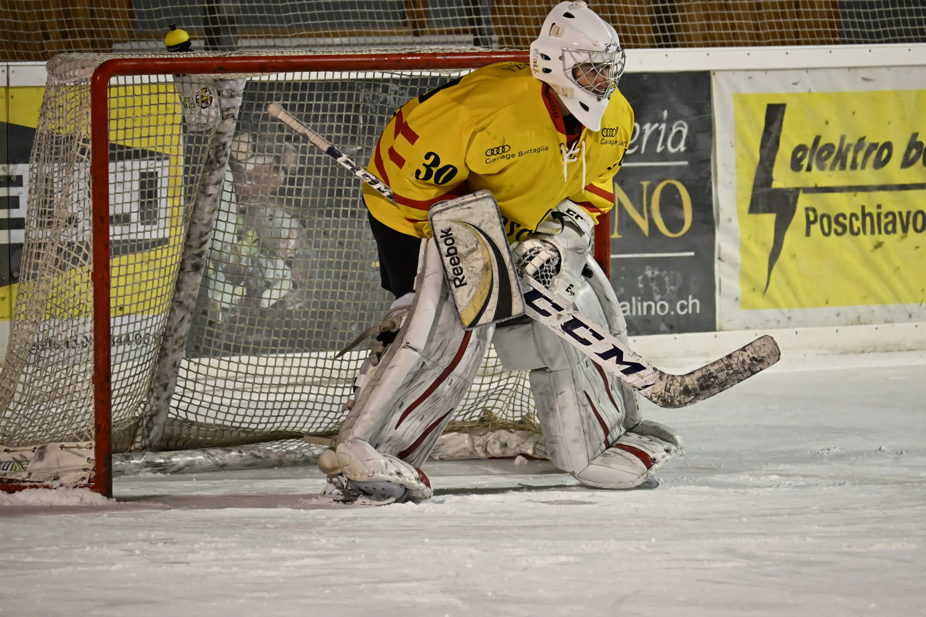 Un portiere di hockey su ghiaccio in divisa gialla sta difendendo la porta. Indossa un casco e protezioni, mentre è in posizione pronta su un ghiaccio ghiacciato