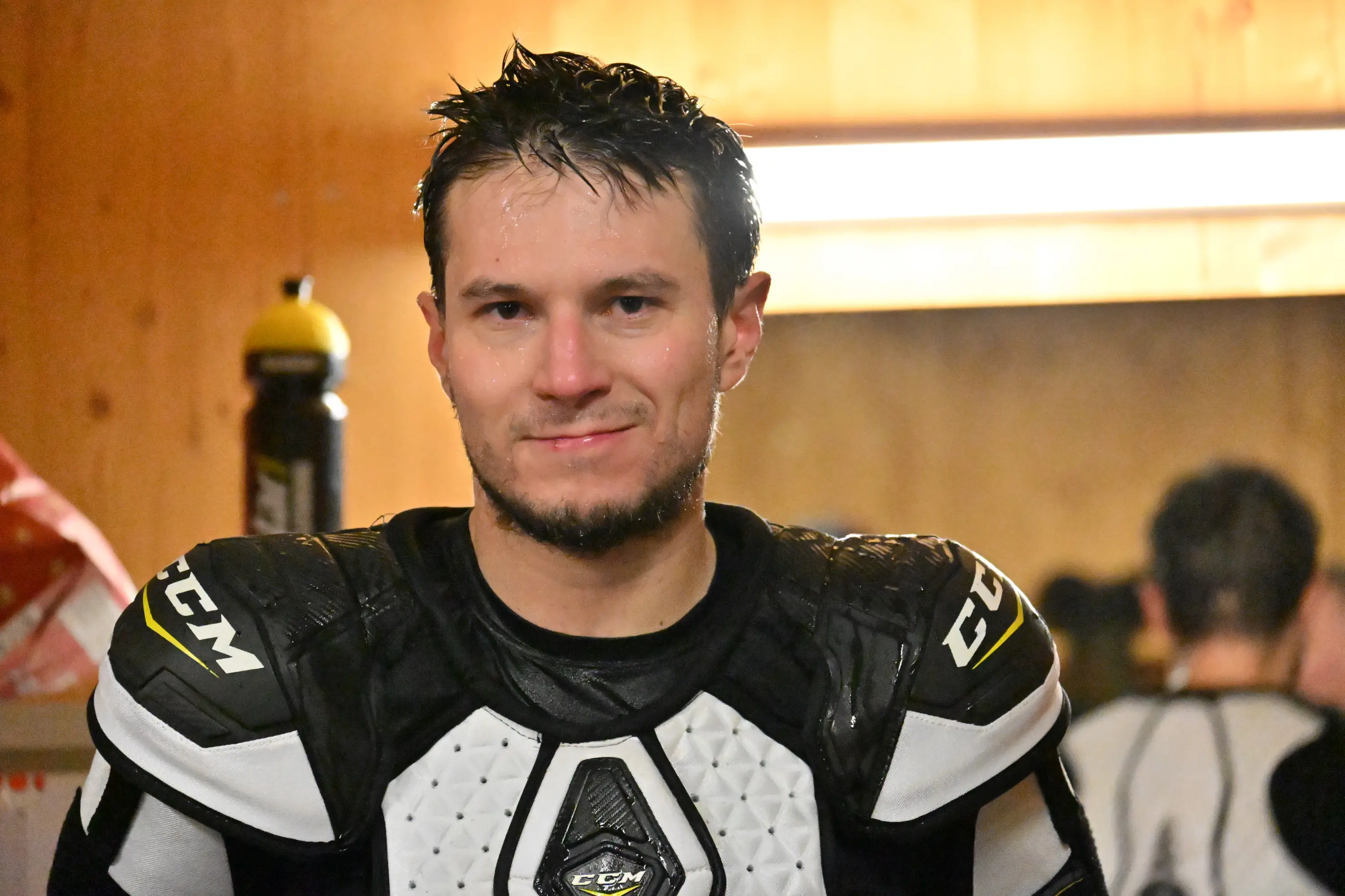 Giovane atleta con attrezzatura da hockey, capelli bagnati e un sorriso, si trova in un ambiente spogliatoio. Sullo sfondo si intravede un'altra persona.